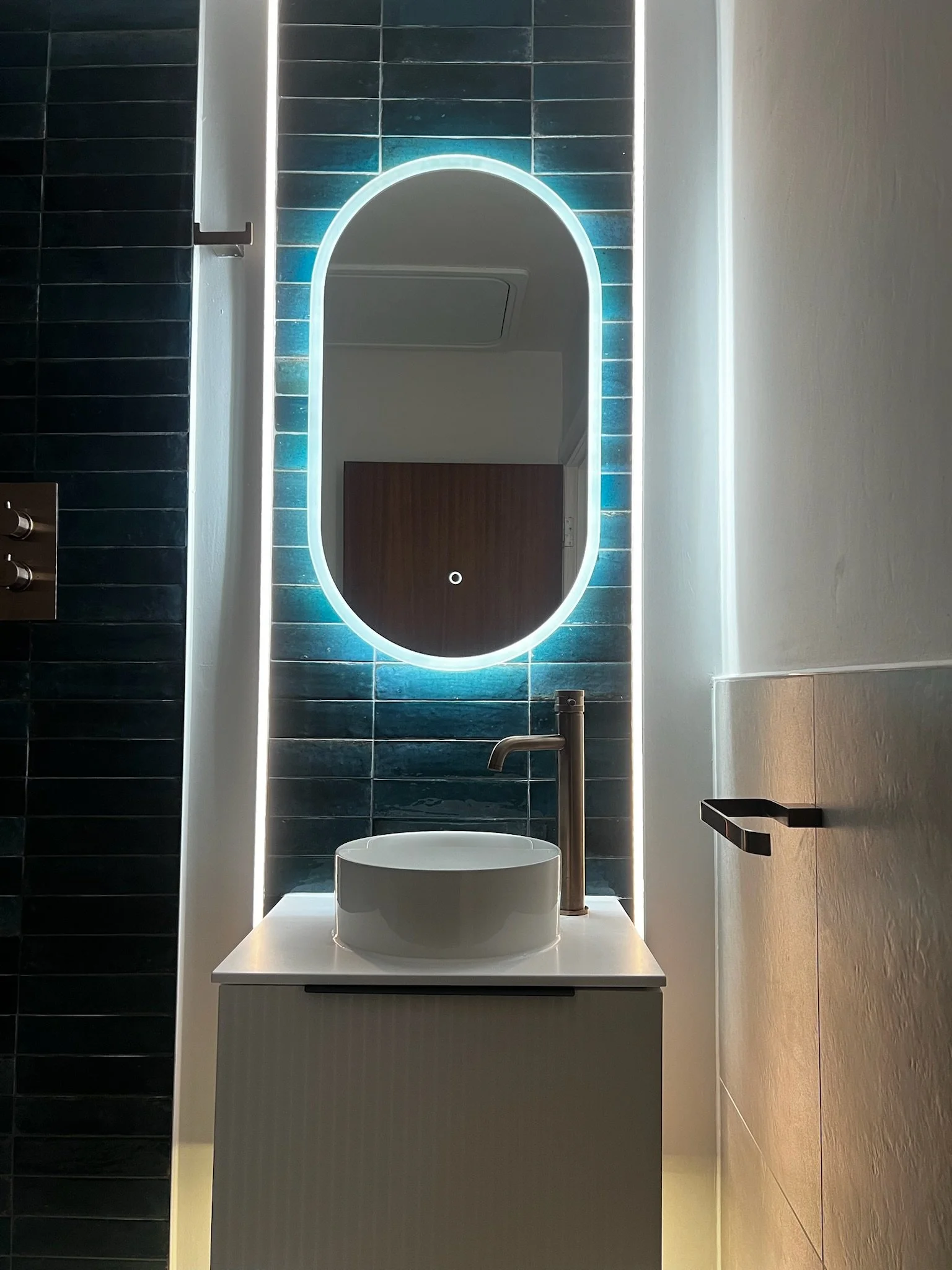 A striking modern bathroom feature in Merseyside showcasing a compact vanity unit with a countertop basin and sleek chrome tap. The space is elevated by a unique backlit mirror with a bold blue glow, set against deep teal tiled walls, creating a dram