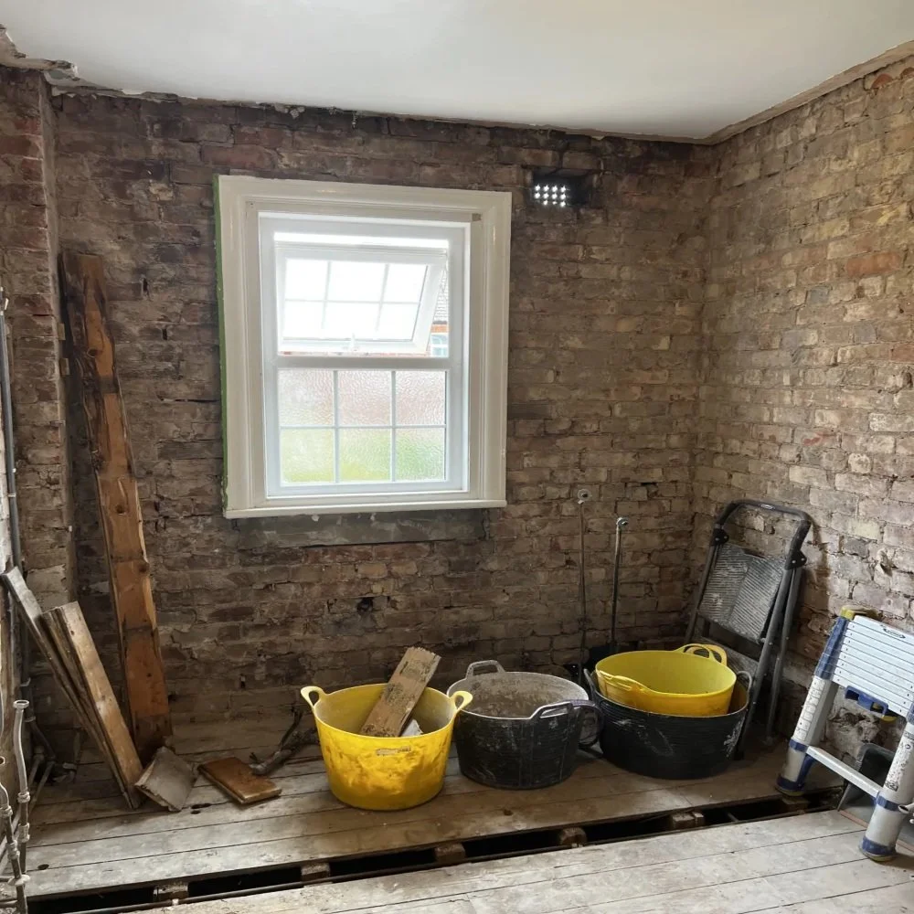 Bathroom renovation in Bebington during strip-out stage showing exposed brick walls, removed flooring, window opening, and buckets with tools and materials ready for installation