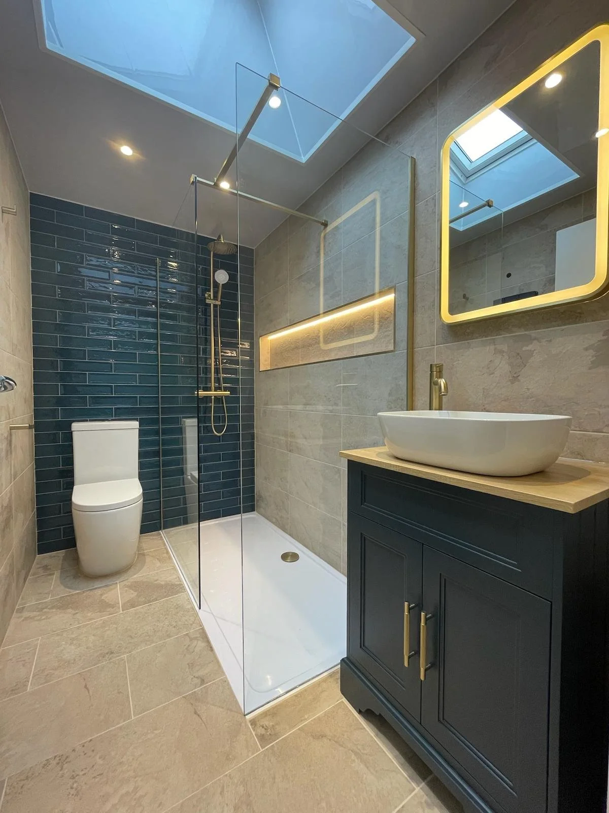 A modern bathroom renovation in Merseyside featuring a walk-in glass shower with striking dark blue tiled walls and sleek gold fixtures. The space includes a contemporary vanity unit with a countertop basin, a softly illuminated mirror, and a skyligh