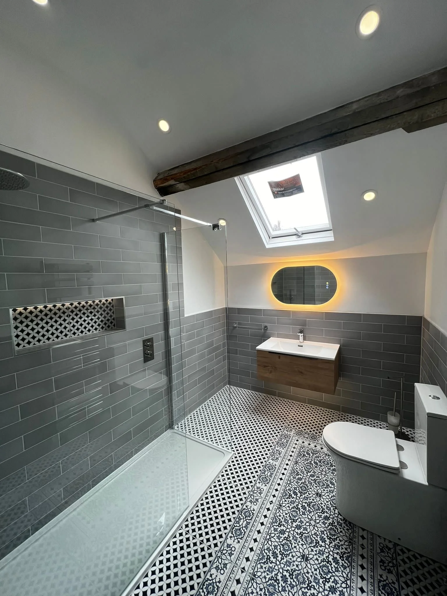 Contemporary bathroom in Merseyside with walk-in shower, grey subway tiles, patterned floor, floating vanity unit, skylight window, and illuminated round mirror.