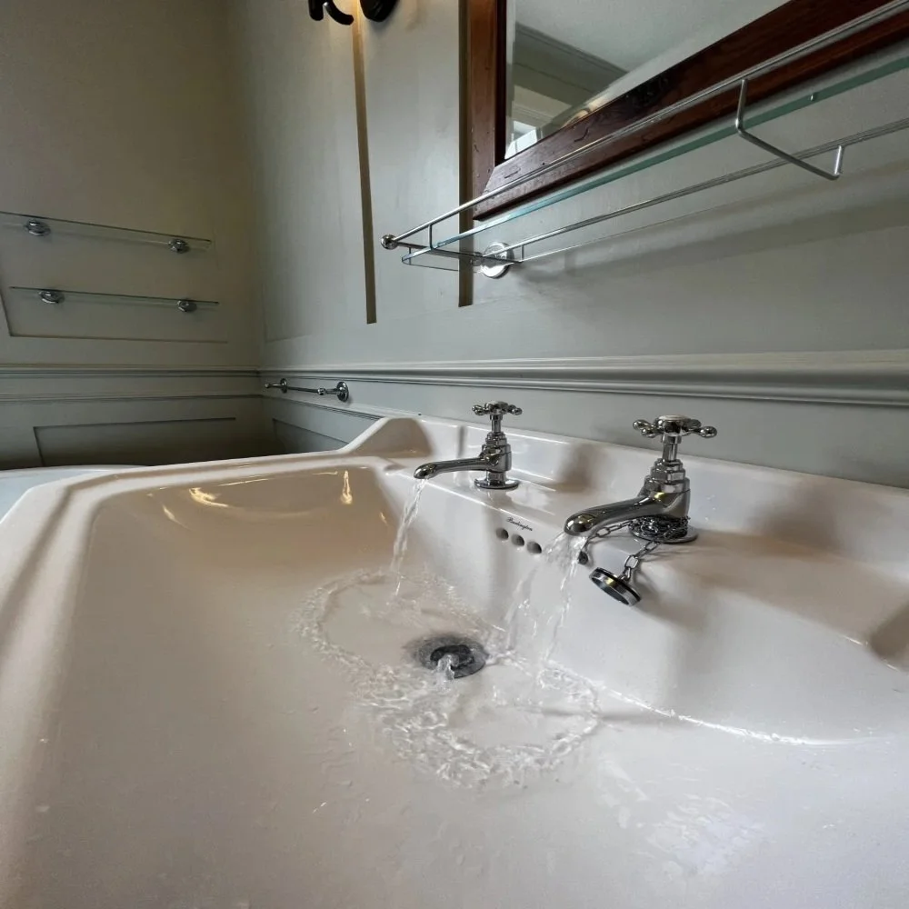 Close-up of a classic bathroom basin in Bebington with chrome hot and cold taps running water, set beneath a mirror and glass shelf against panelled walls