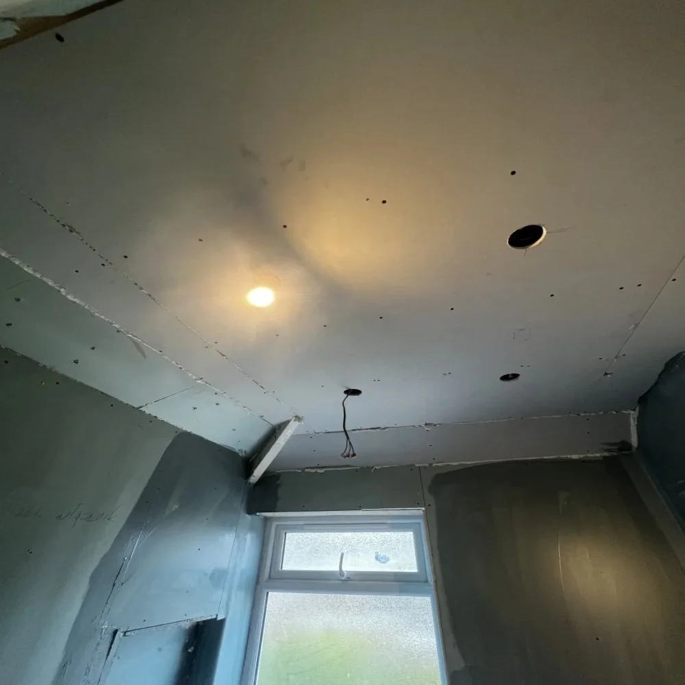 Bathroom renovation in Aigburth showing newly installed plasterboard ceiling with cut-outs for downlights, exposed wiring, and partially finished walls around window