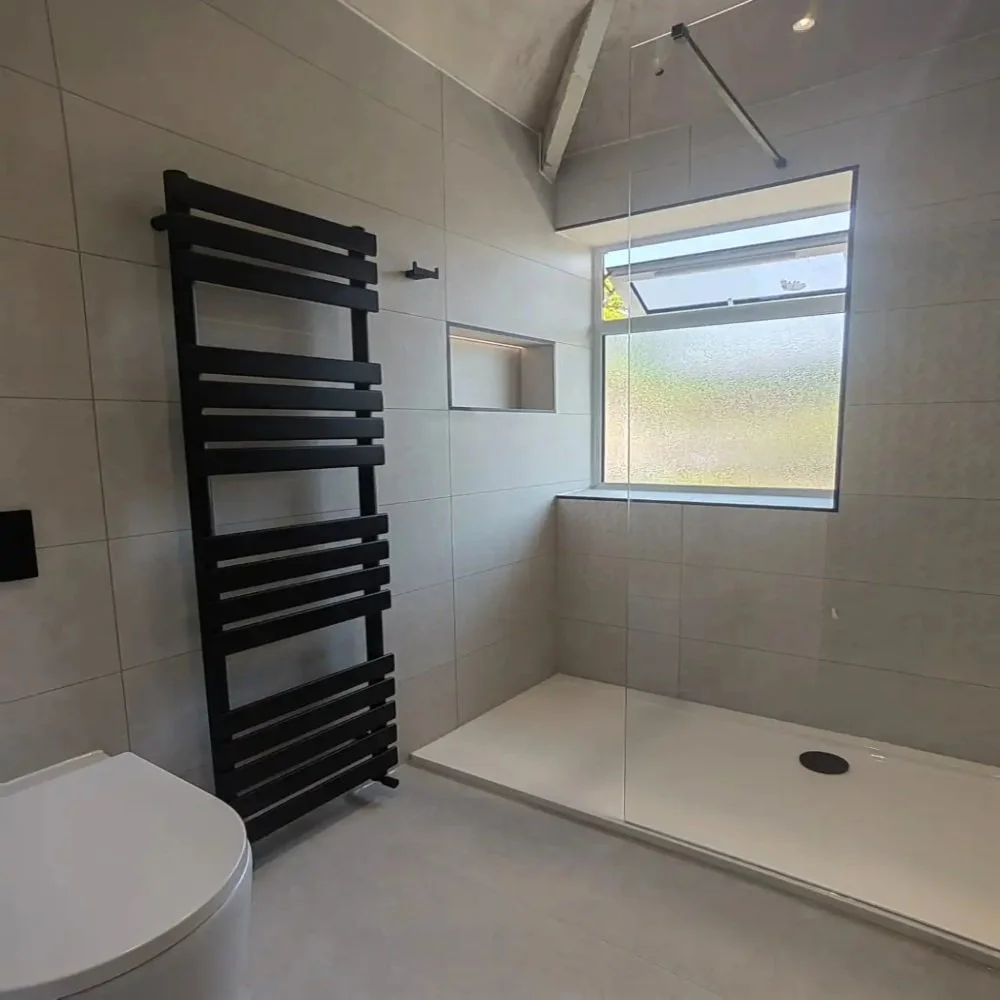 Modern bathroom in Aigburth featuring walk-in shower with glass screen, recessed wall niche, black towel radiator, and large light tiles with frosted window above