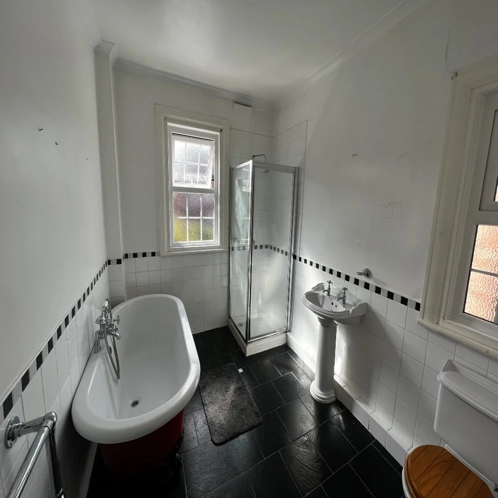 Bright traditional bathroom in Bebington featuring a freestanding roll-top bath, glass shower enclosure, pedestal sink, black floor tiles, and white tiled walls with black border detailing