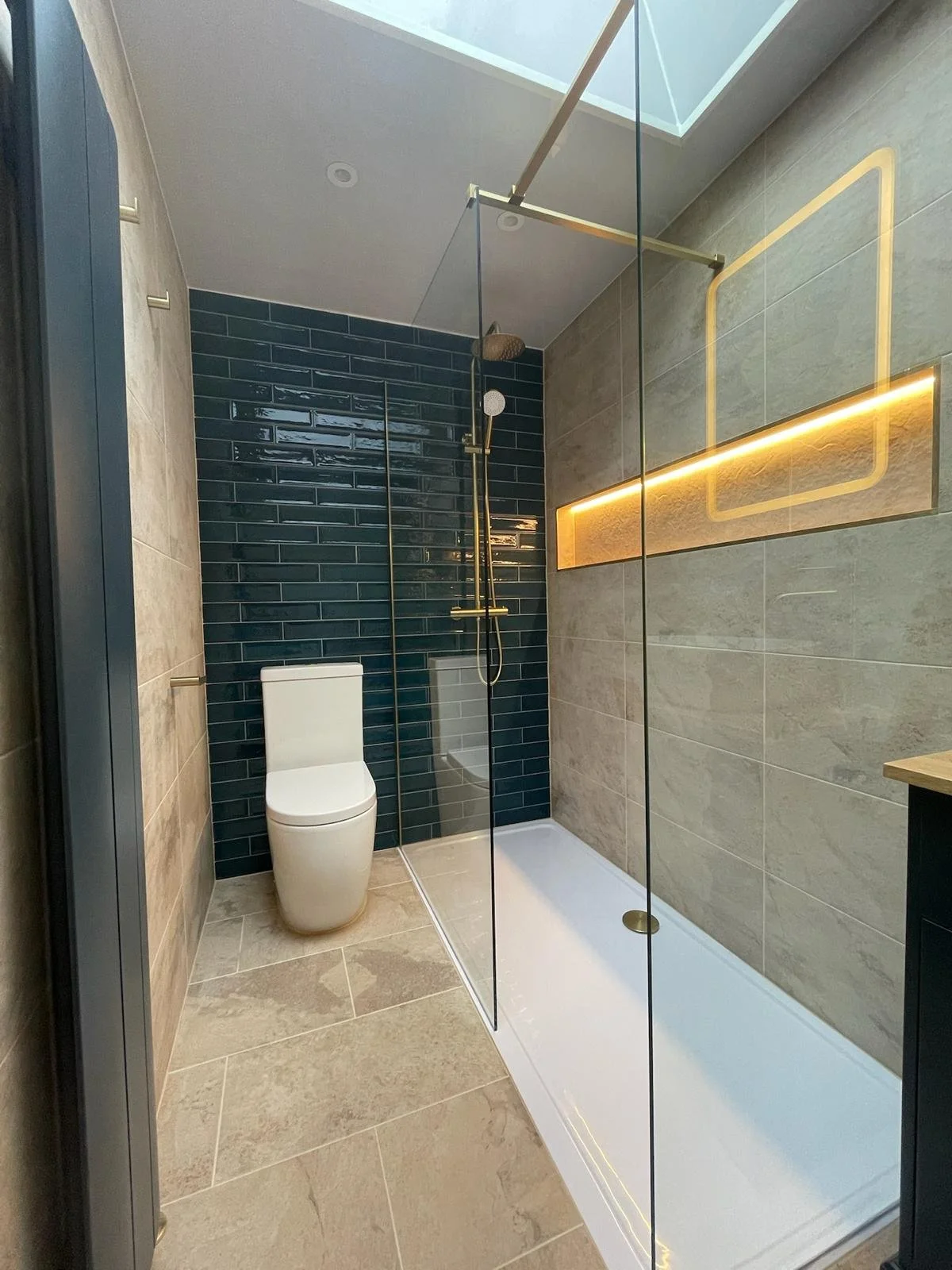 A sleek and modern bathroom renovation in Merseyside featuring a walk-in glass shower with elegant gold fixtures and a rainfall showerhead. The space is highlighted by deep blue tiled walls within the shower area, contrasted with neutral stone-effect