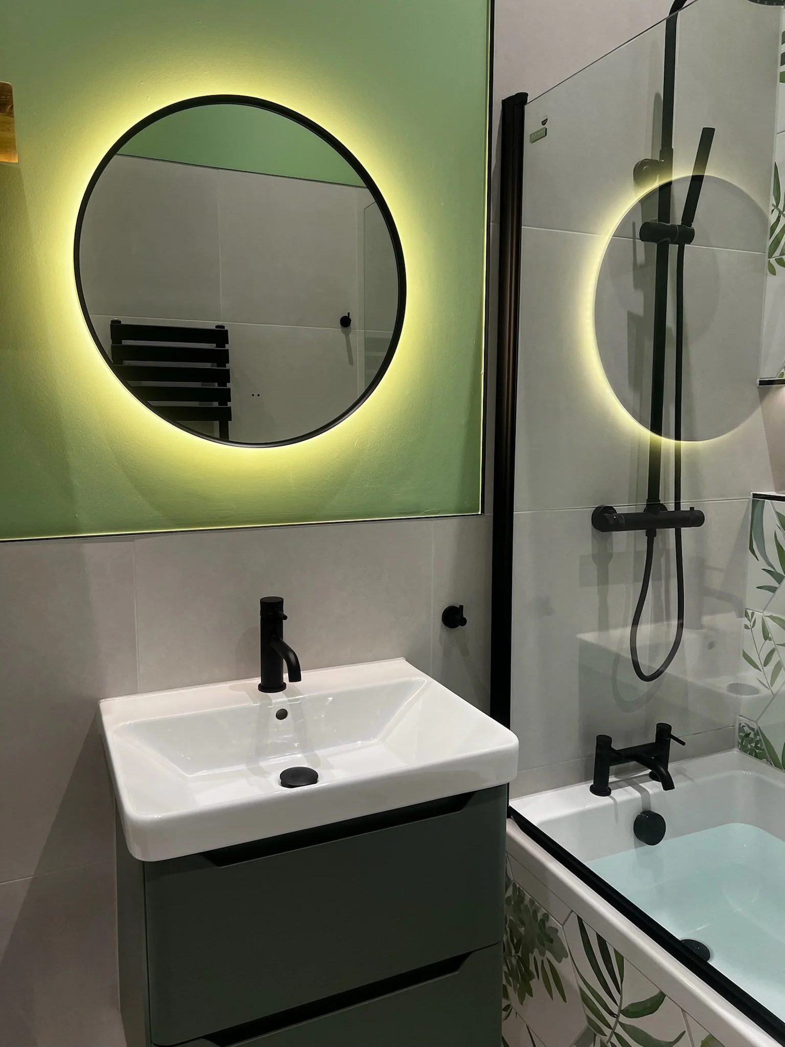 A modern bathroom renovation in Merseyside featuring a compact wall-mounted vanity unit with an integrated basin and matte black tap. The space is enhanced by a round backlit mirror with a soft glow, paired with a bath and overhead shower area with m