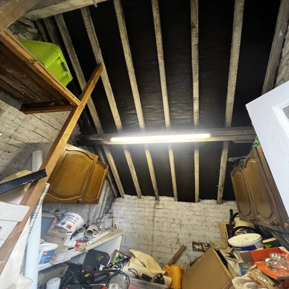 Loft area with exposed timber rafters and brick walls, filled with stored items and old cabinets, being cleared during a bathroom renovation in Bebington