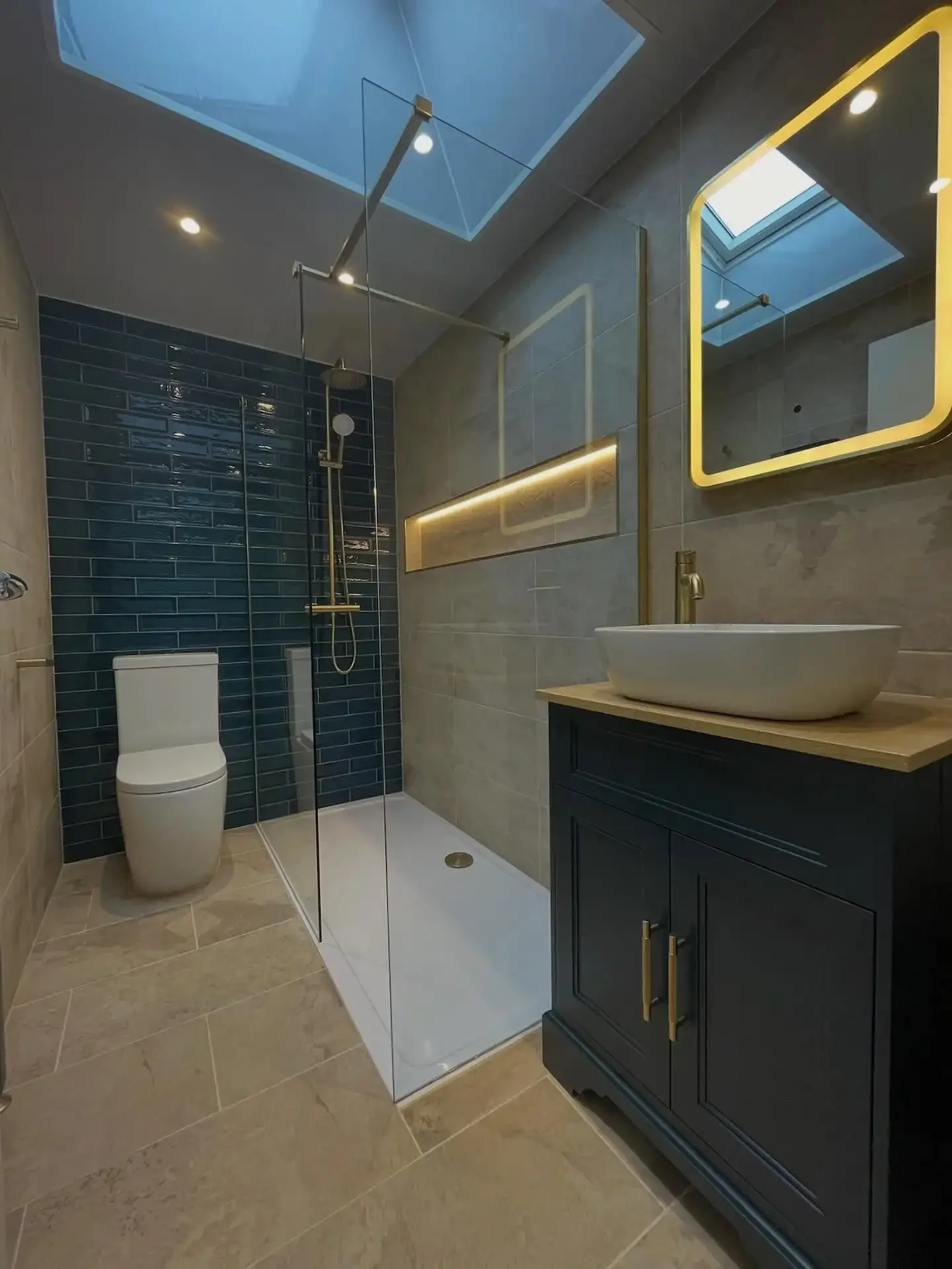 A stylish and contemporary bathroom renovation in Merseyside featuring a walk-in glass shower with deep blue tiled walls and elegant gold fixtures. The space includes a vanity unit with a countertop basin, a softly illuminated mirror, and a skylight 