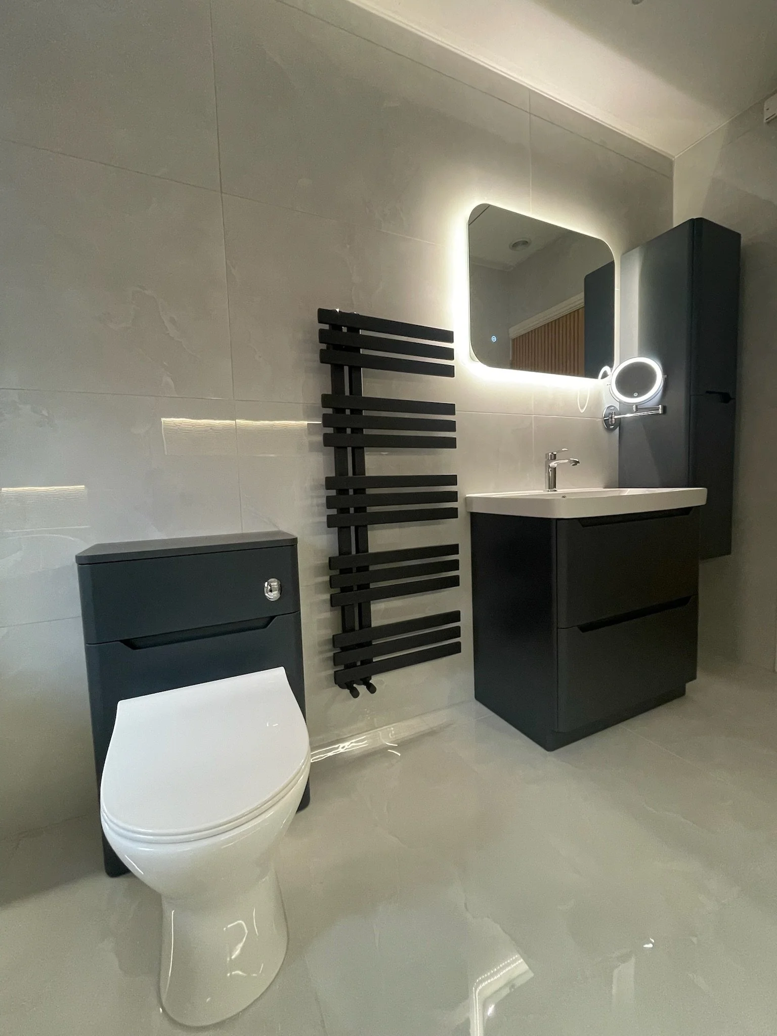 A sleek and modern bathroom renovation in Merseyside featuring a compact vanity unit with an integrated basin and a contemporary WC. The space is enhanced by a softly illuminated mirror, a black heated towel rail, and large neutral tiles, creating a 