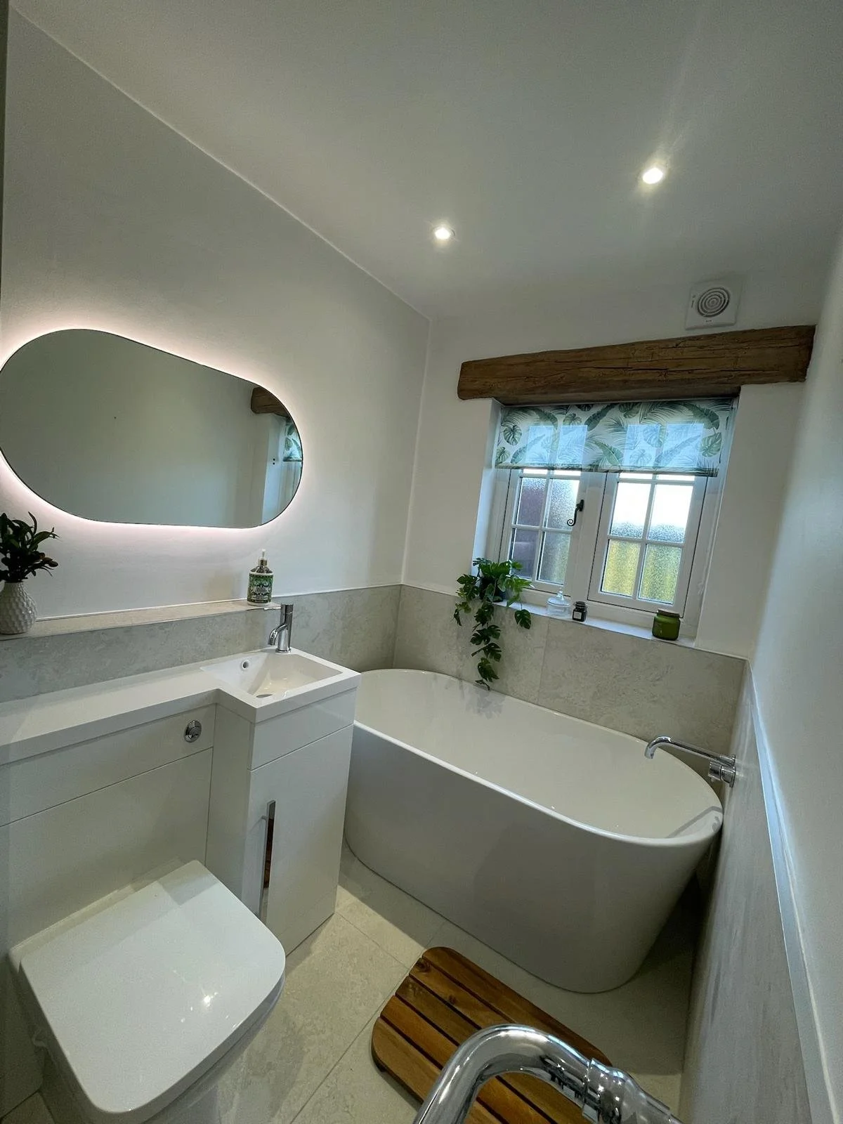 Stylish small bathroom in Merseyside with freestanding bath, wall-hung vanity, soft LED mirror lighting, natural wood accents, and a bright window with plants for a fresh modern feel.