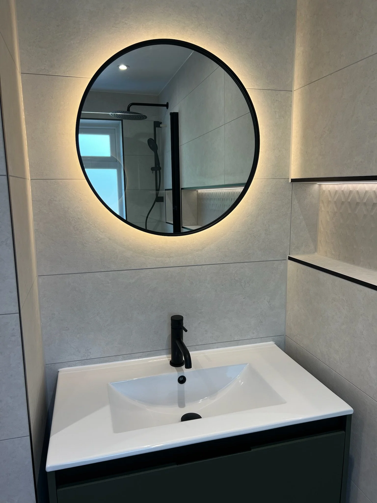 A sleek and minimalist bathroom renovation in Merseyside featuring a wall-mounted vanity unit with a modern integrated basin and matte black tap. The space is enhanced by a round mirror and clean-lined neutral tiles, with a recessed shelf adding both
