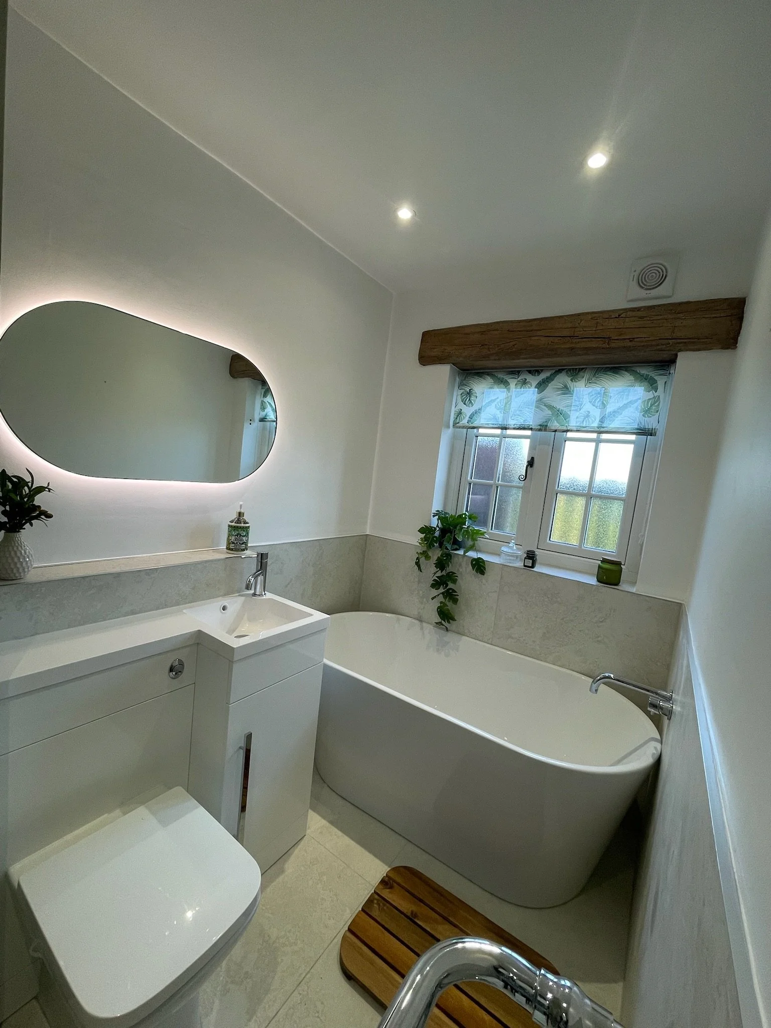 A bright and elegant bathroom renovation in Merseyside featuring a sleek freestanding bath positioned beneath a window, allowing plenty of natural light to fill the space. The design includes a wall-mounted vanity unit with an integrated basin, a sof