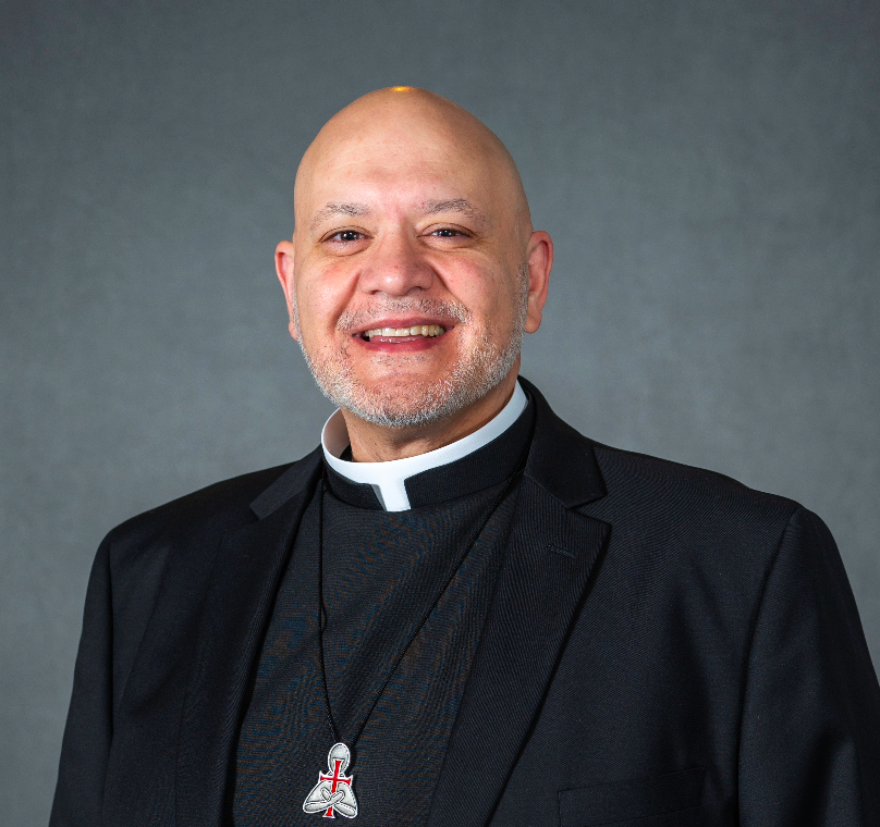 A smiling middle-aged man wearing a priest's clerical collar and black suit, with a cross pendant necklace, standing against a plain gray background.