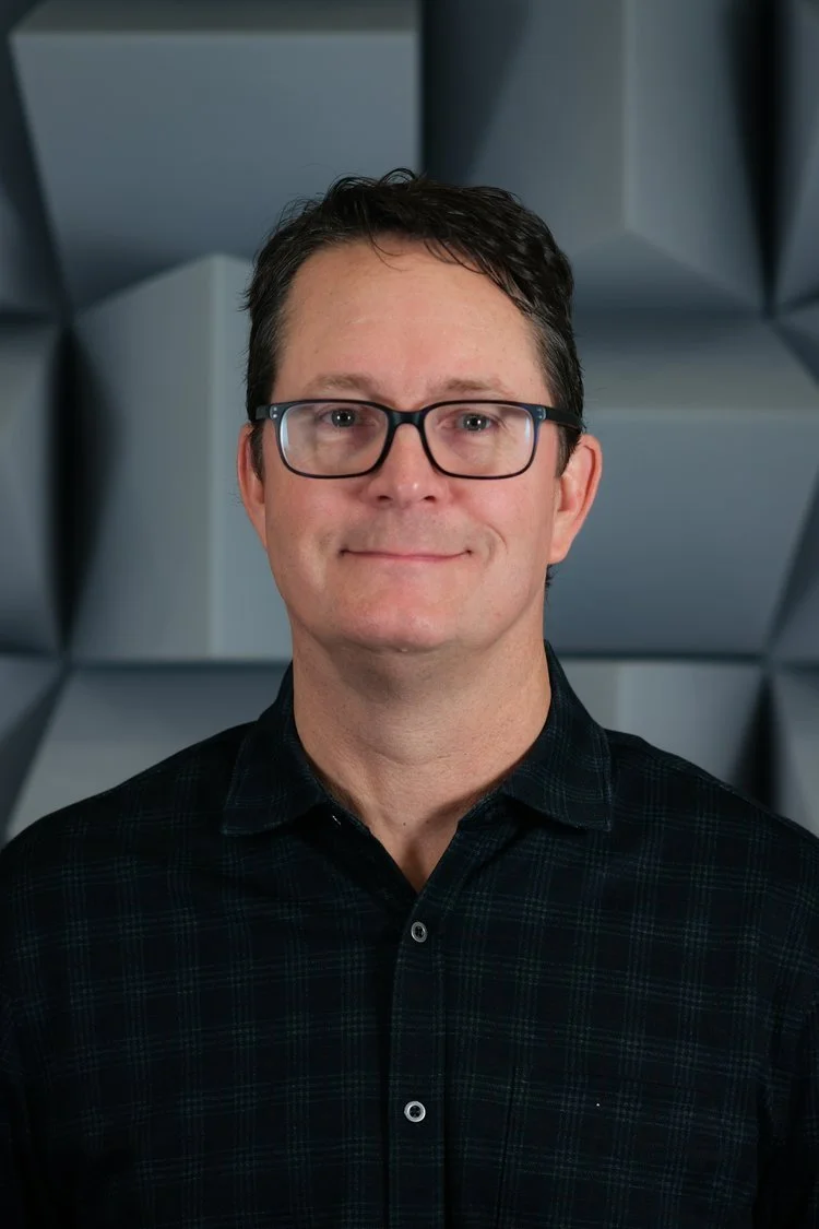 A man with short dark hair, glasses, and a slight smile wearing a dark plaid shirt in front of a geometric gray background.