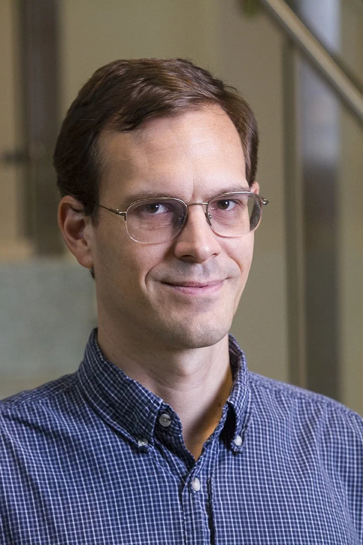 Portrait of a man with brown hair and glasses, wearing a blue checkered shirt, standing indoors against a blurred background.