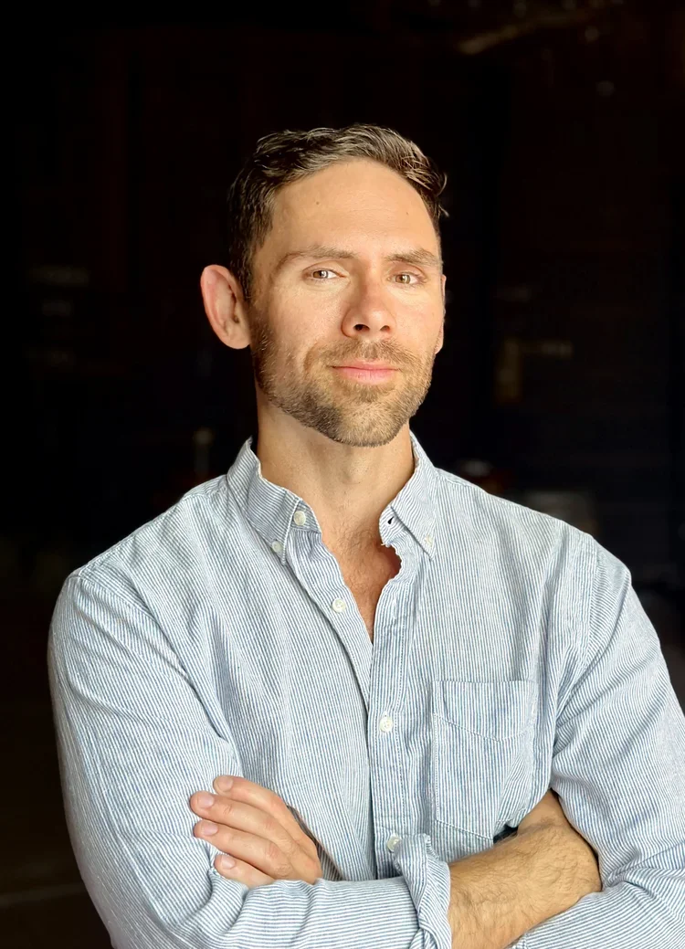 A man with short dark hair and a beard, wearing a light blue striped button-up shirt, standing with arms crossed against a dark background.