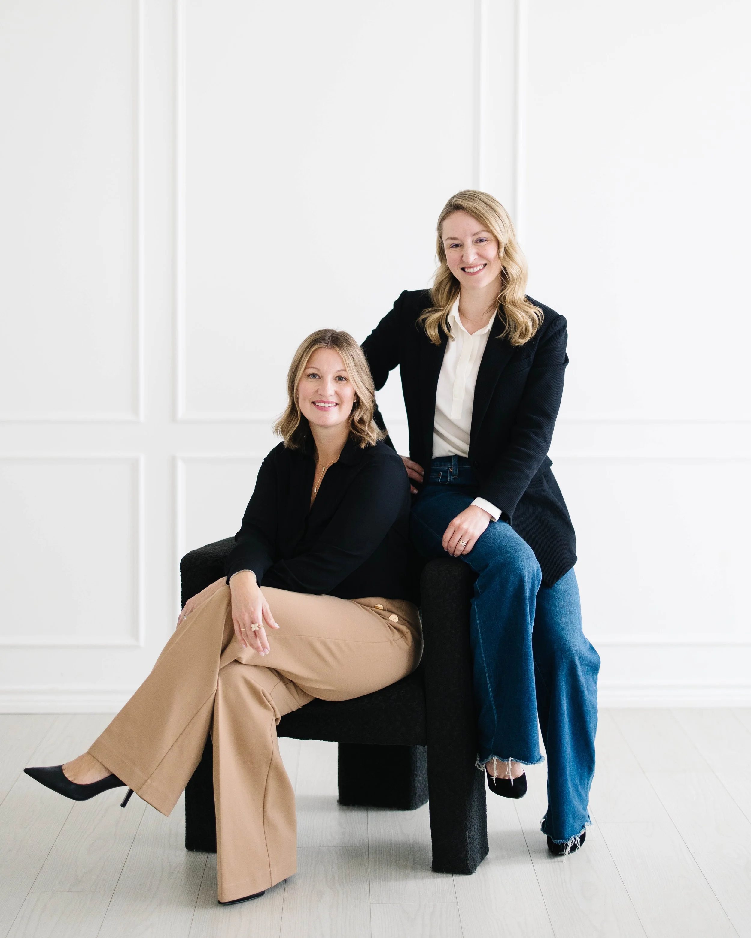 Claire Peacock and Aimee Bradley dressed in business attire pose together in a white room, with one sitting and the other standing beside her, smiling at the camera.