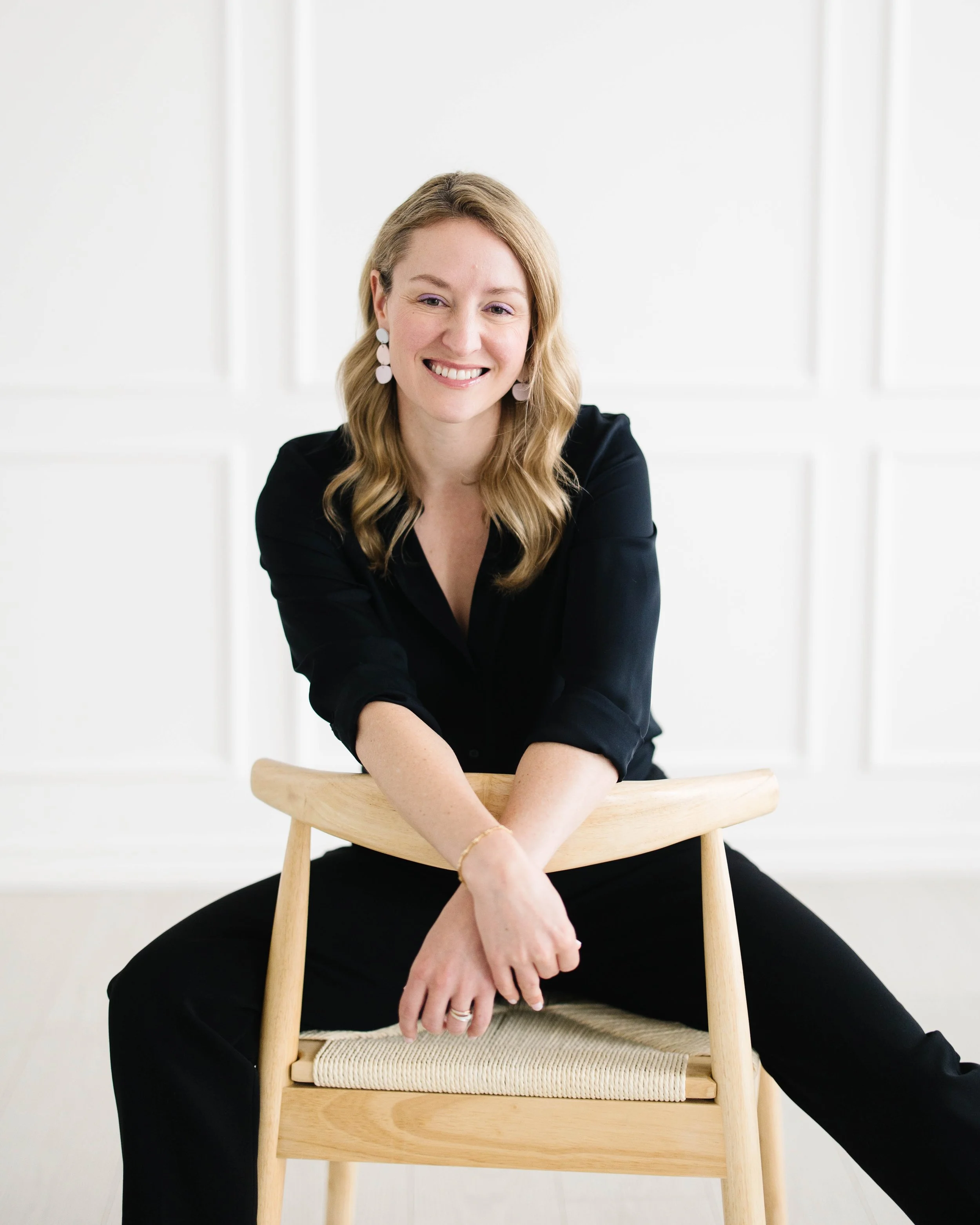 Claire Peacock with blonde hair smiling, wearing a black top and black pants, sitting behind a wooden chair with a woven seat, in a bright, minimalistic room.