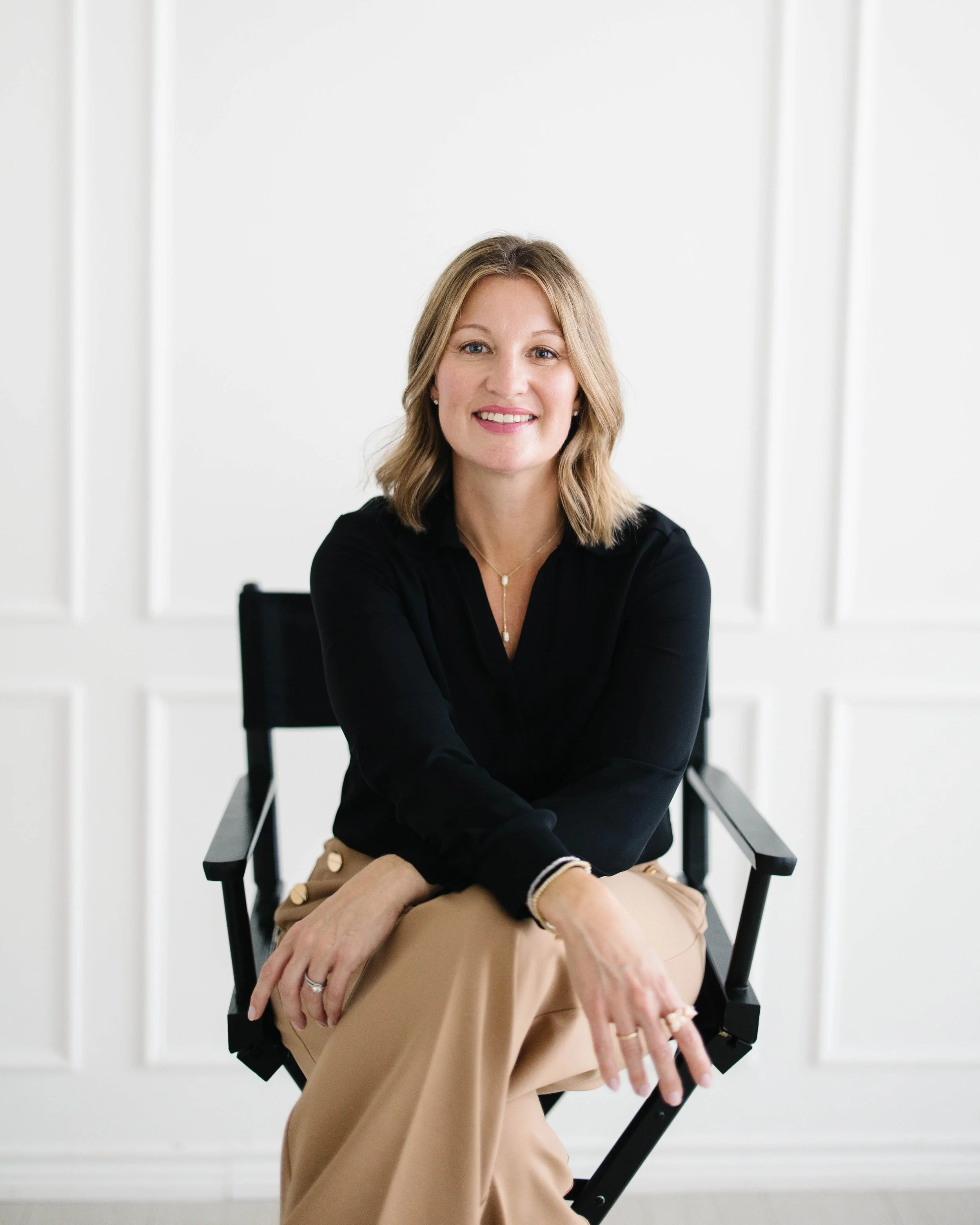 Aimee Bradley with blonde hair, wearing a black top and beige pants, sitting on a black director's chair against a white background, with white paneled walls.