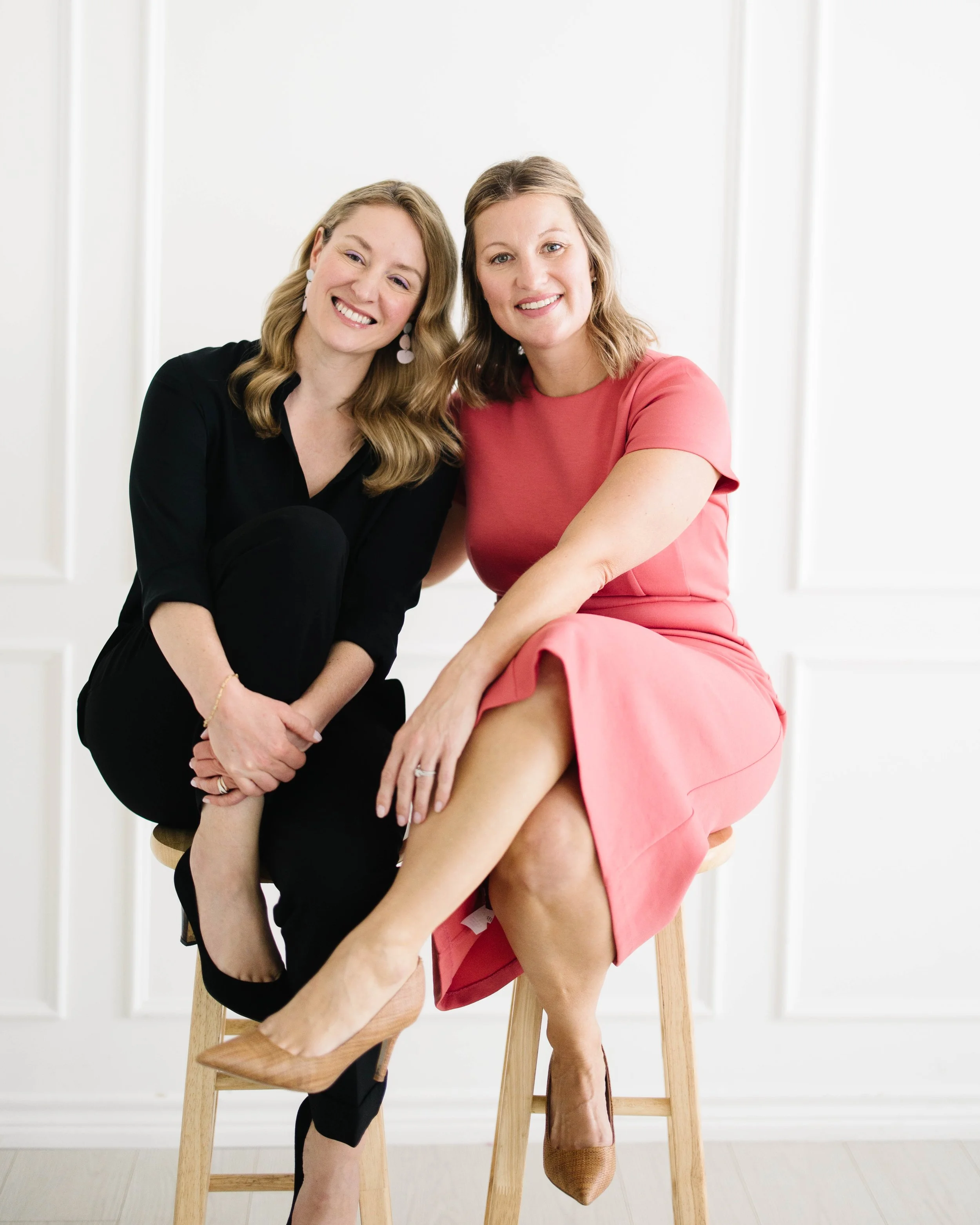 Claire Peacock and Aimee Bradley sitting on wooden stools in front of a white panel wall, smiling at the camera, one wearing a black top and the other in a pink dress with one leg crossed over the other.