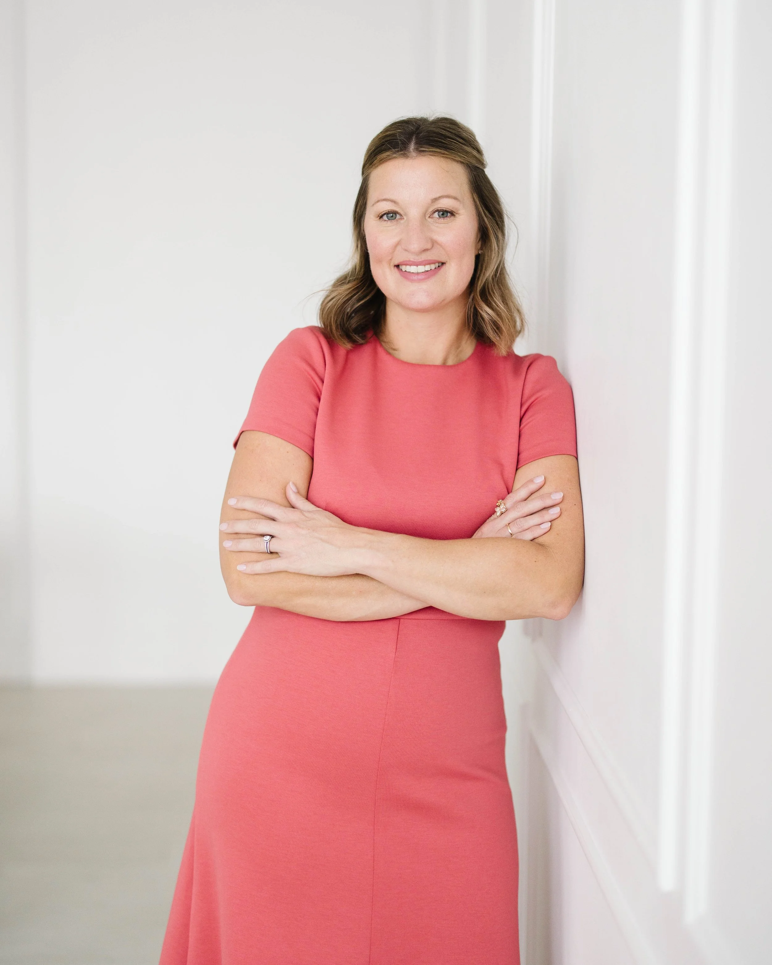 Aimee Bradley, co-founder of Candere Creative Strategy wearing a pink dress, standing indoors with crossed arms, smiling at the camera.