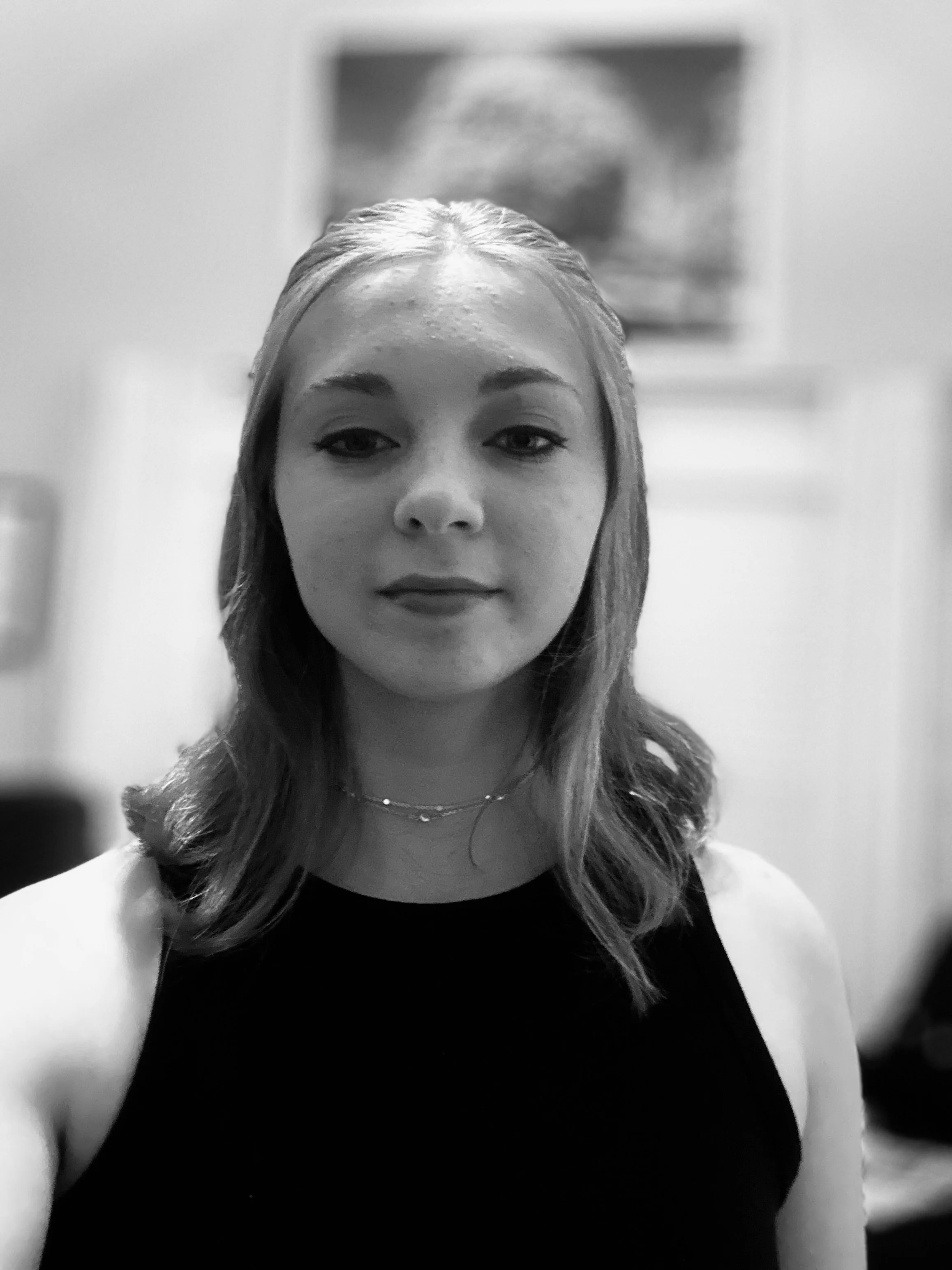 A woman with shoulder-length hair, wearing a sleeveless top and a delicate necklace, taking a selfie indoors with a blurred background.