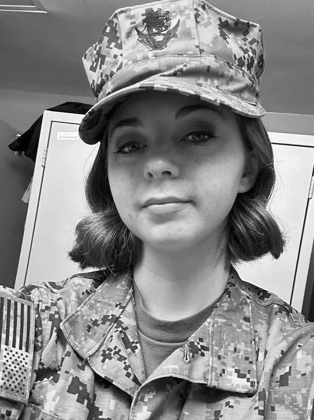 A female sailor in uniform and camouflage hat taking a selfie in a locker room.