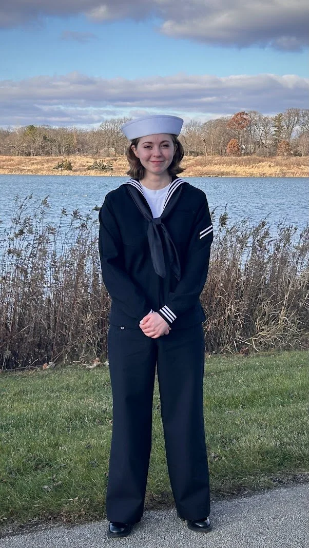Young woman in a navy sailor uniform standing outdoors near a lake with dry grass and trees in the background.