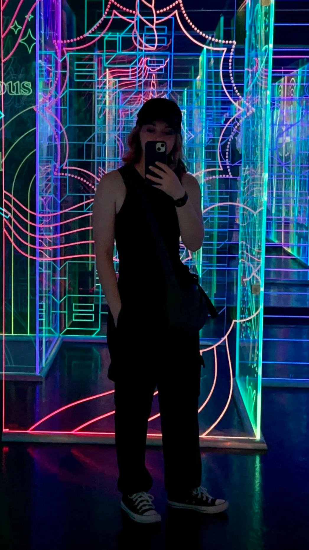 Person taking a mirror selfie in front of neon light art installation with colorful, glowing geometric and floral patterns.