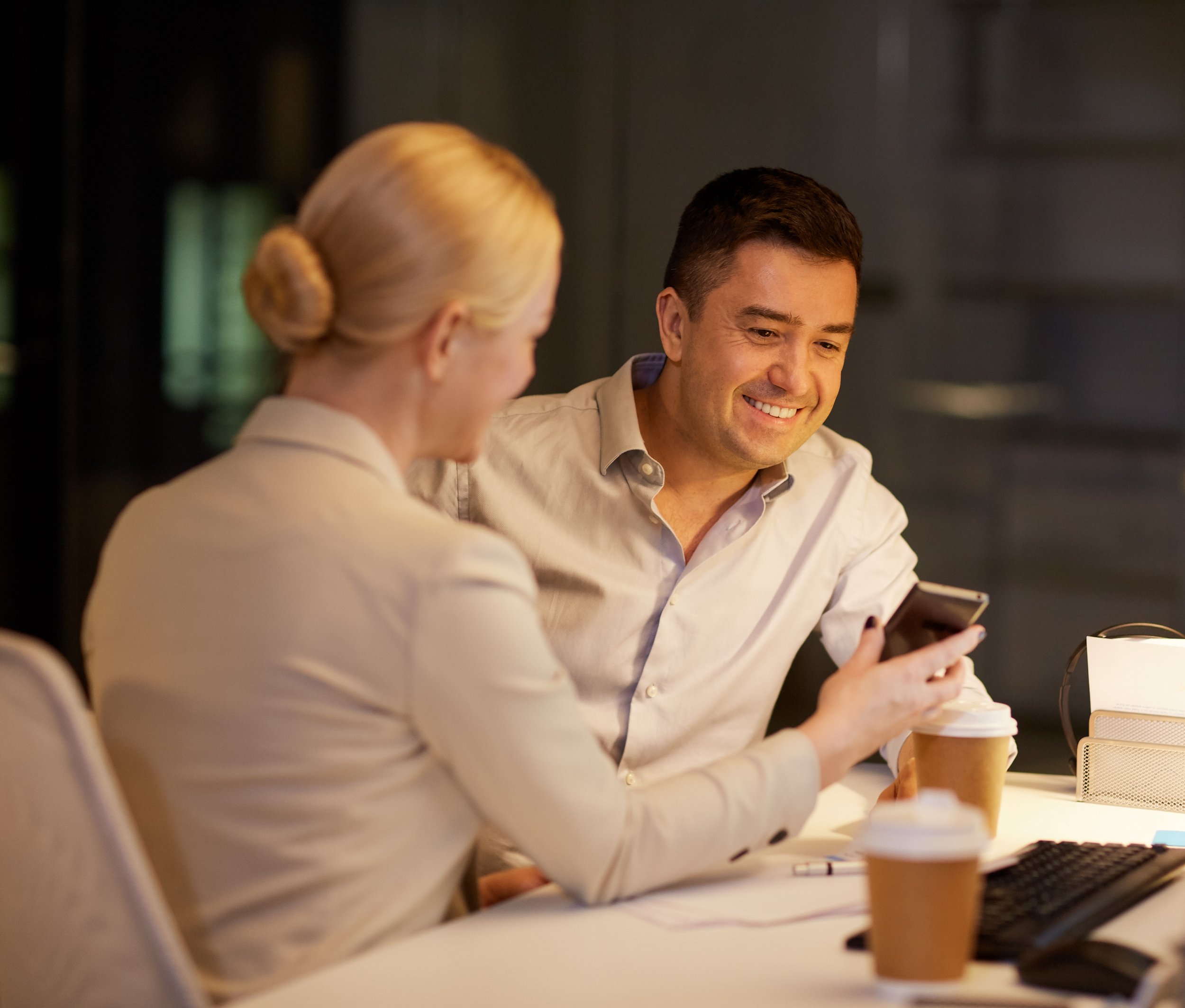 Two colleagues, a man and a woman, sitting at a table in a professional setting, using a smartphone, with coffee cups on the table.