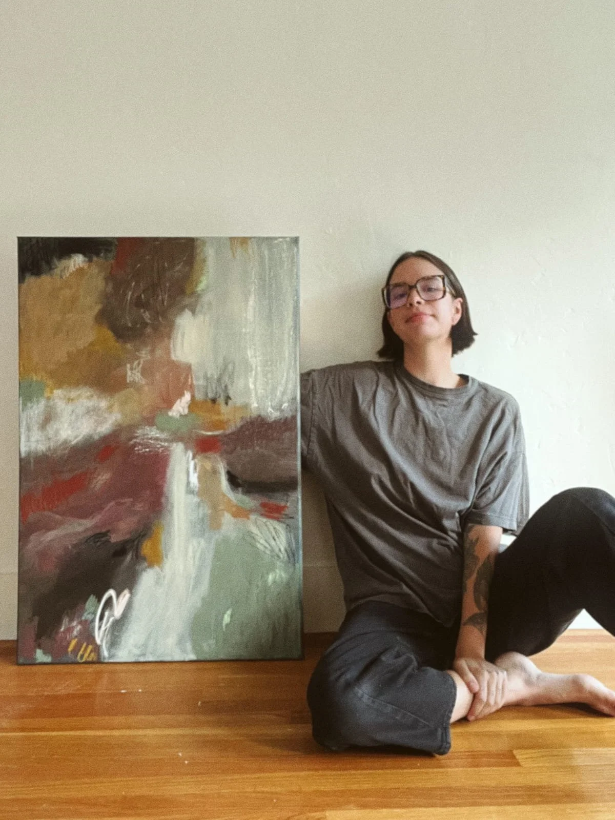 A person sitting on the floor next to a colorful abstract painting, wearing glasses and a gray t-shirt, with a relaxed pose and a slight smile.