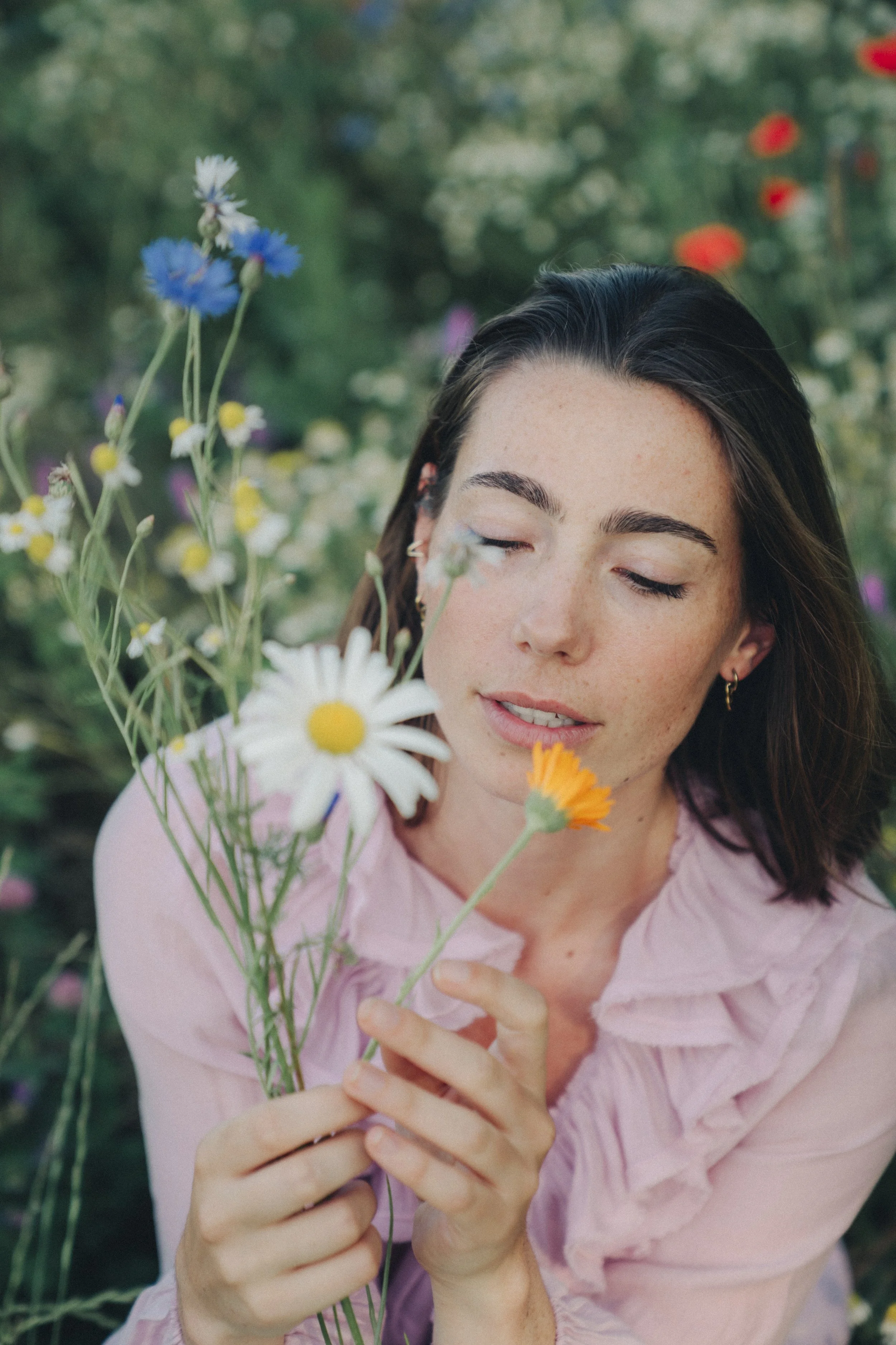 A woman with dark hair and freckles, wearing a light pink shirt, holding a bouquet of wildflowers with her eyes closed in a lush garden.