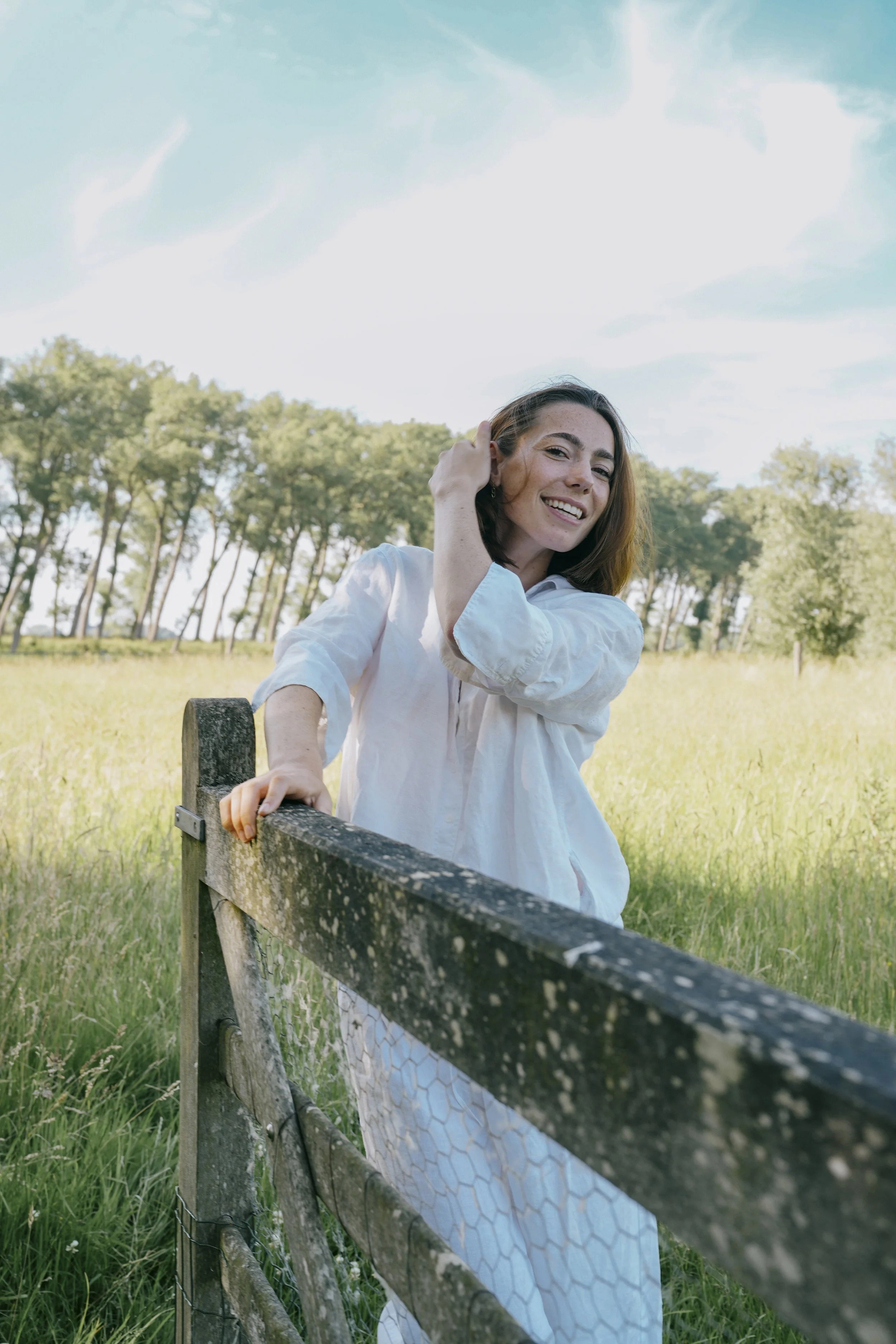 A woman with shoulder-length brown hair, wearing a white shirt, is smiling and leaning on a rustic wooden fence in a green field with tall grass and trees in the background, under a partly cloudy sky.