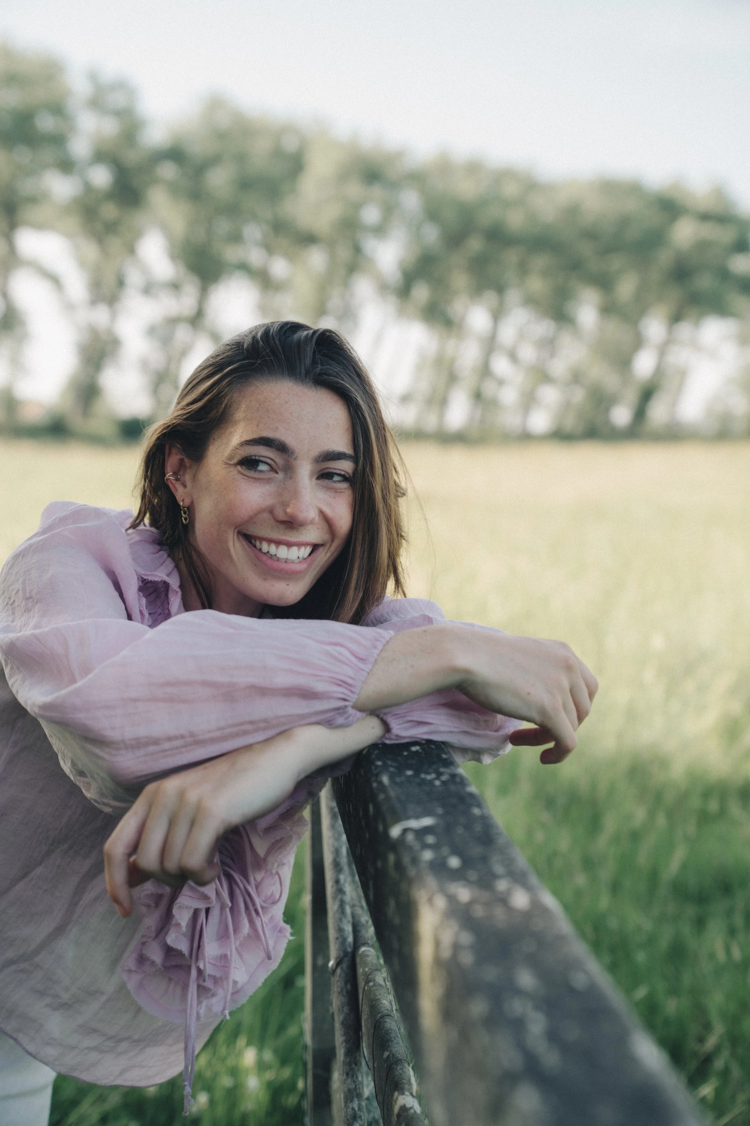 A young woman with long brown hair, wearing a pink blouse and earrings, is leaning on a black metal railing outdoors on a grassy field with blurry trees in the background. She is smiling and looking at the camera.