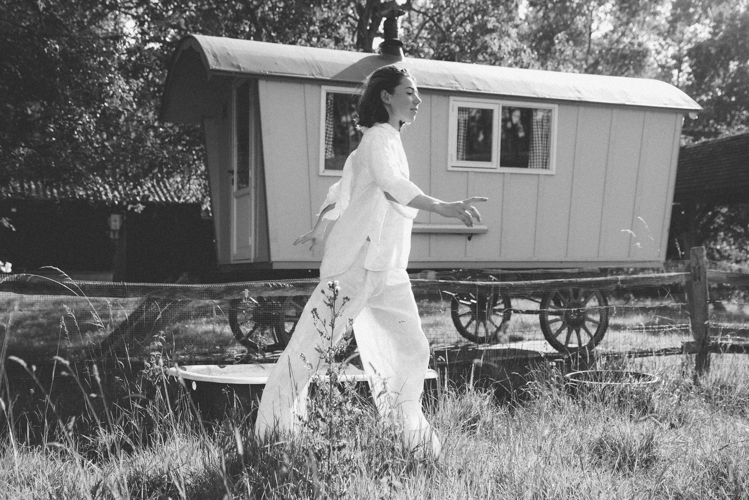A woman dressed in white walking outdoors with trees and a small white shed in the background.