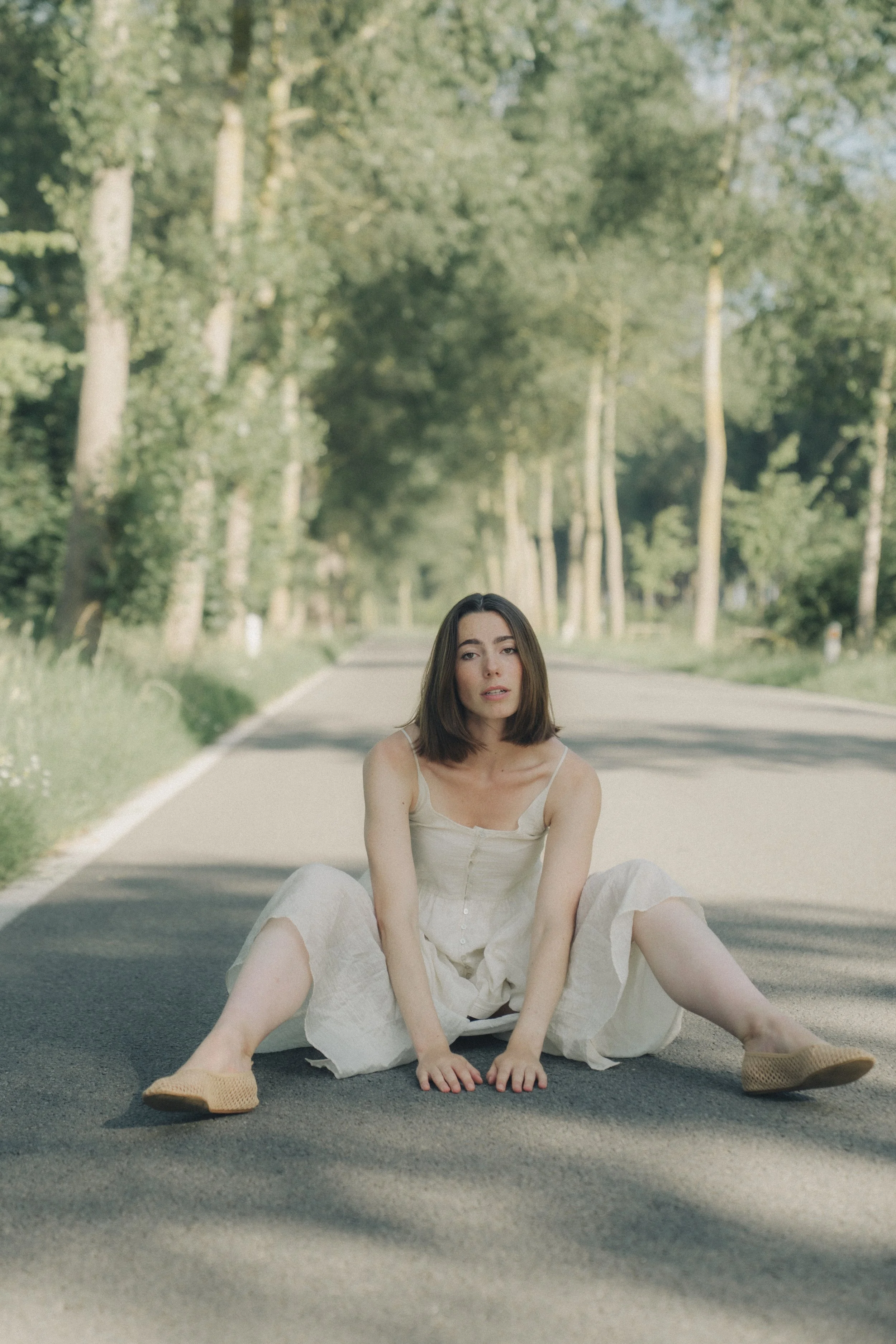 A young woman sitting on the middle of a deserted road in a rural area with trees on both sides, wearing a white dress and beige shoes, with a neutral expression.