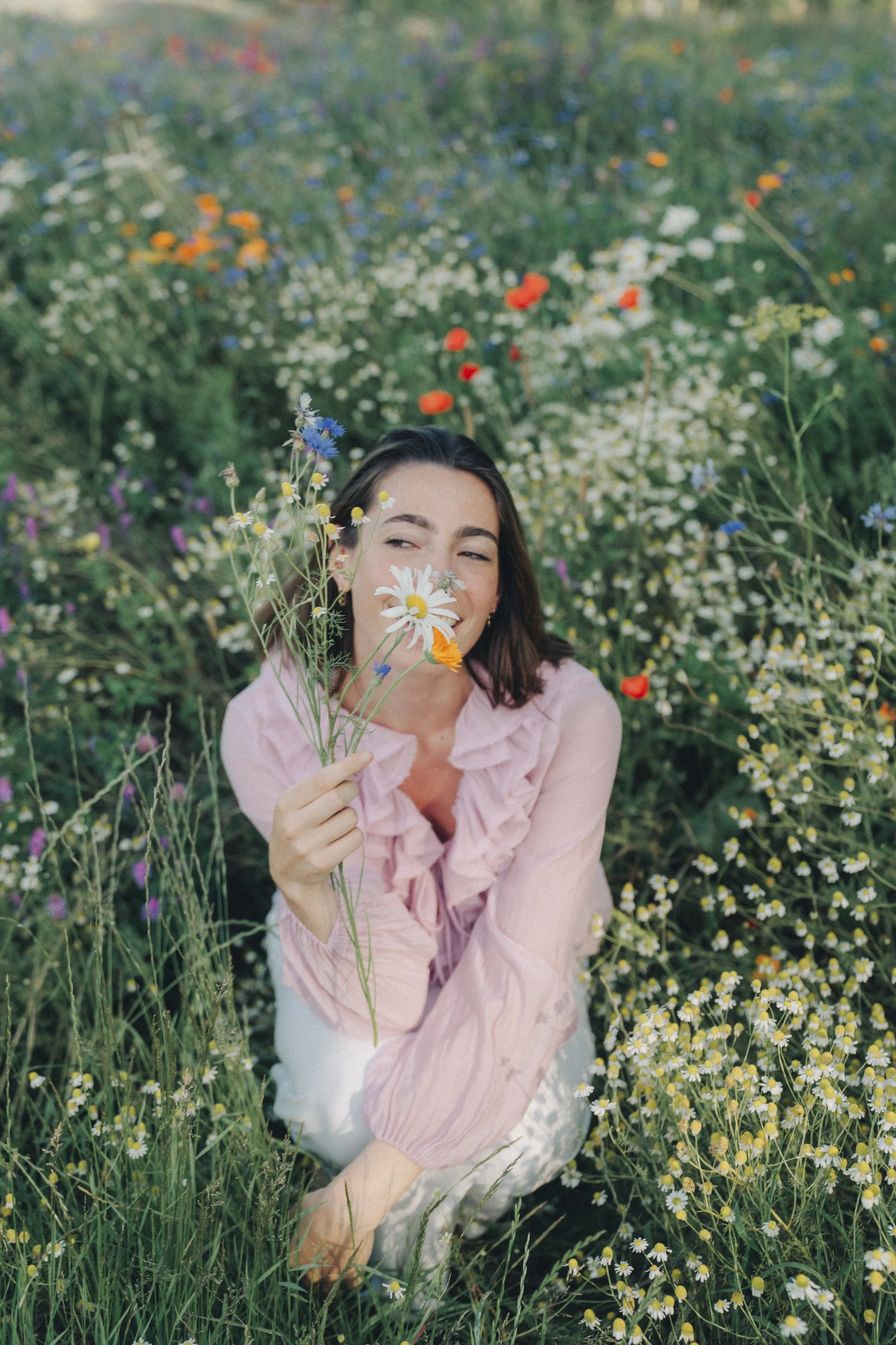 A woman with dark hair and light skin holding a small bouquet of wildflowers, smiling and squinting among wildflowers in a green meadow.