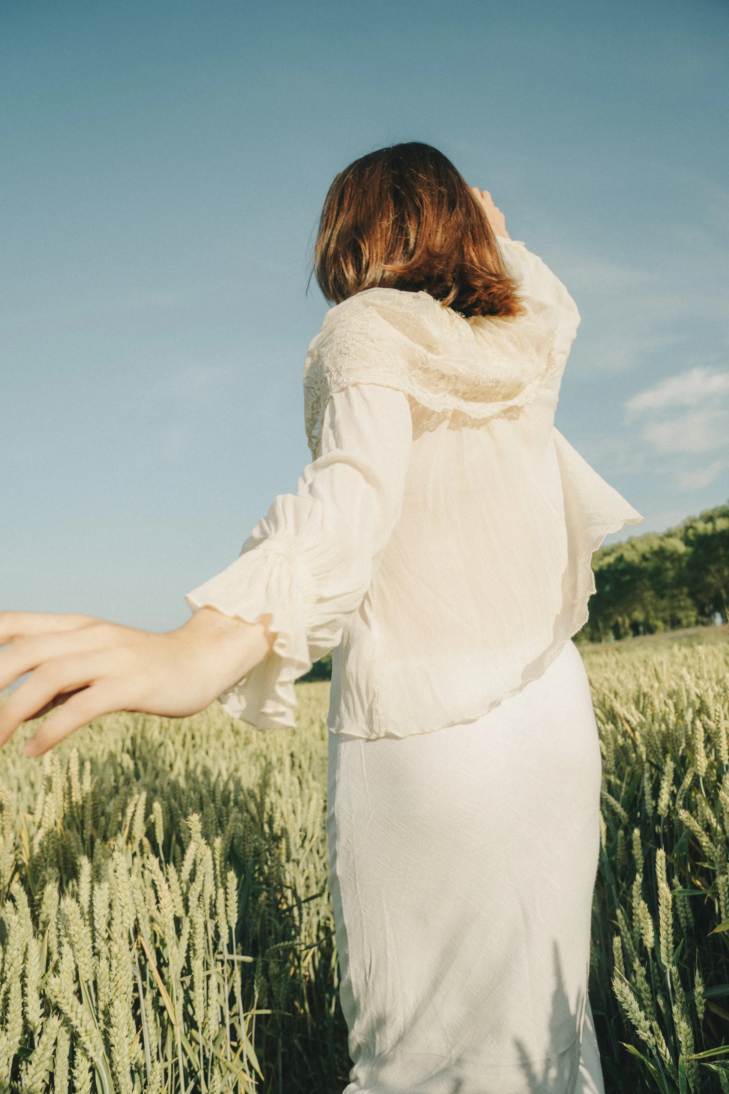 A person with brown hair is holding hands with someone outside in a field of wheat under a clear blue sky.