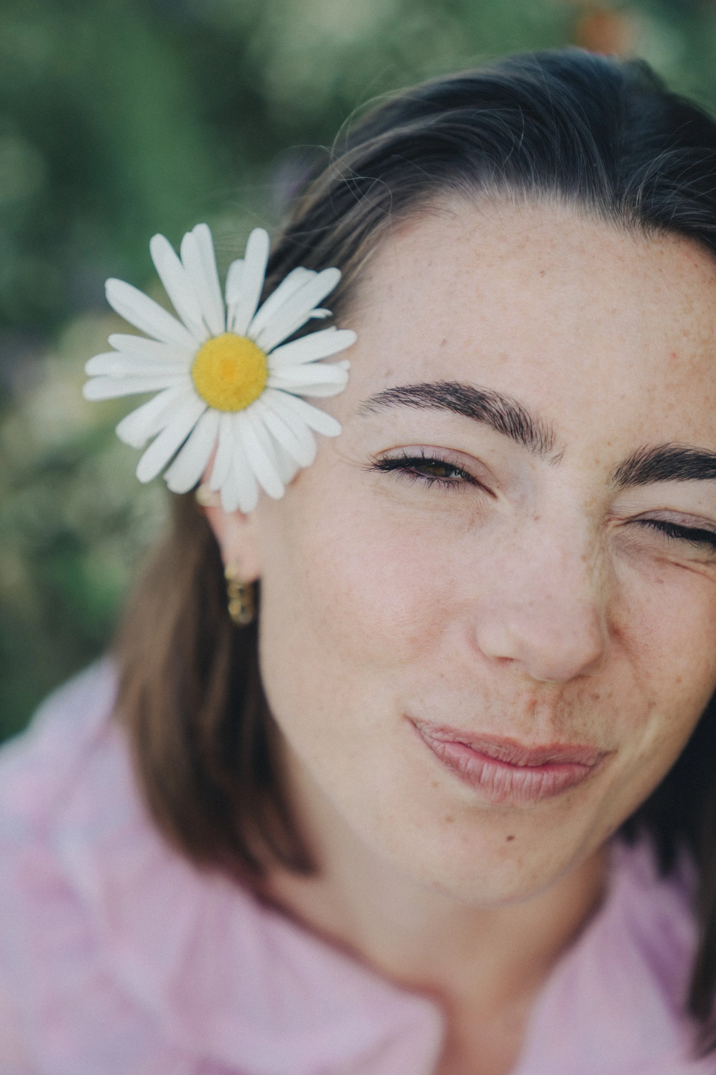Close-up of a woman's face with a daisy flower tucked behind her ear, smiling softly with the background blurred green foliage.