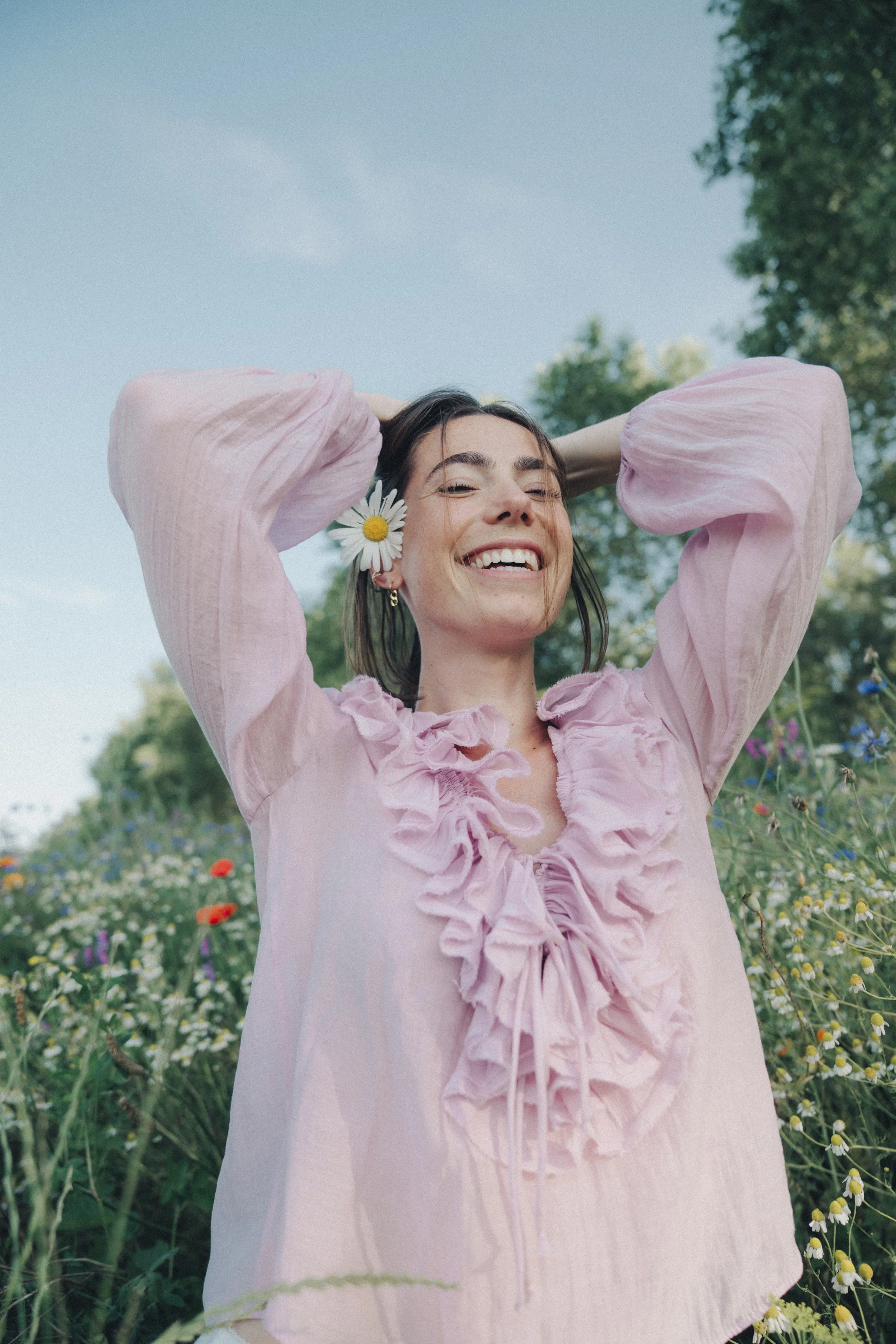 Smiling woman with a daisy next to her face, wearing a light pink dress with ruffled details, standing in a field of wildflowers under a blue sky.