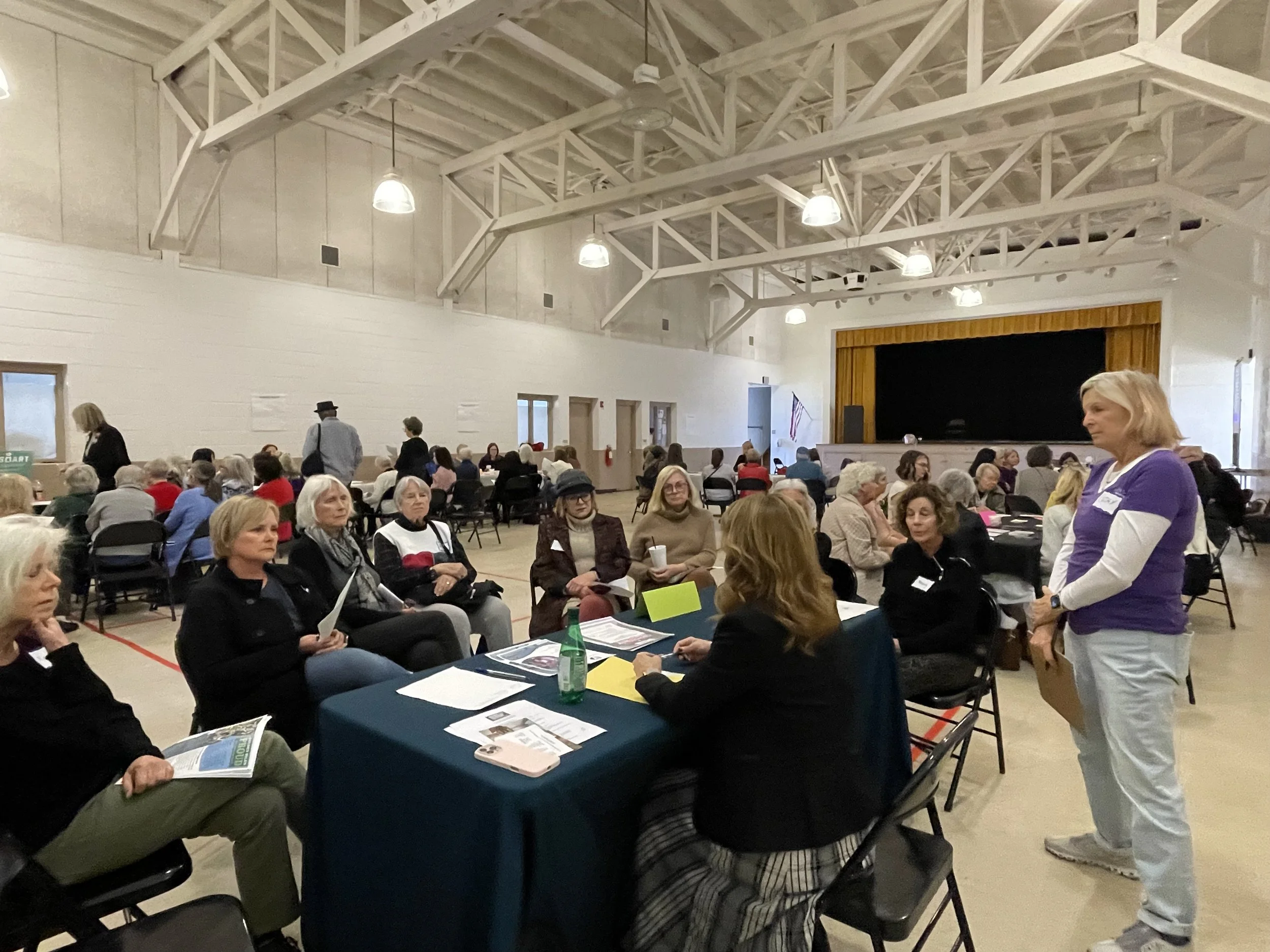 A large gathering of people seated at tables inside a community hall with a stage and a yellow curtain at the back. Some people are talking, reading pamphlets, or sitting quietly. The hall has a high ceiling with white exposed beams, and several hanging lights.