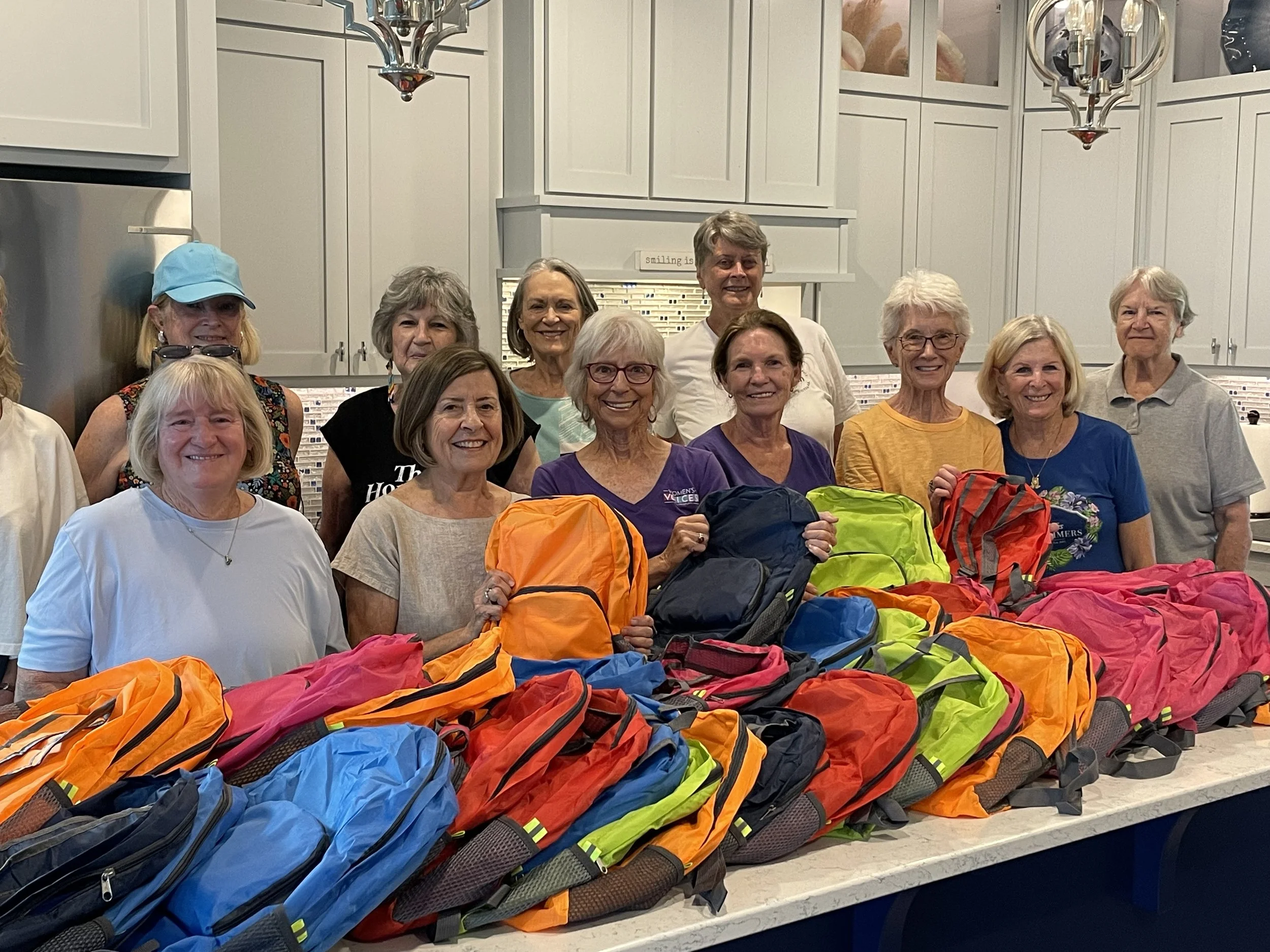 Group of smiling women gathered in a kitchen, holding colorful backpacks, with more backpacks on the counter in front of them.