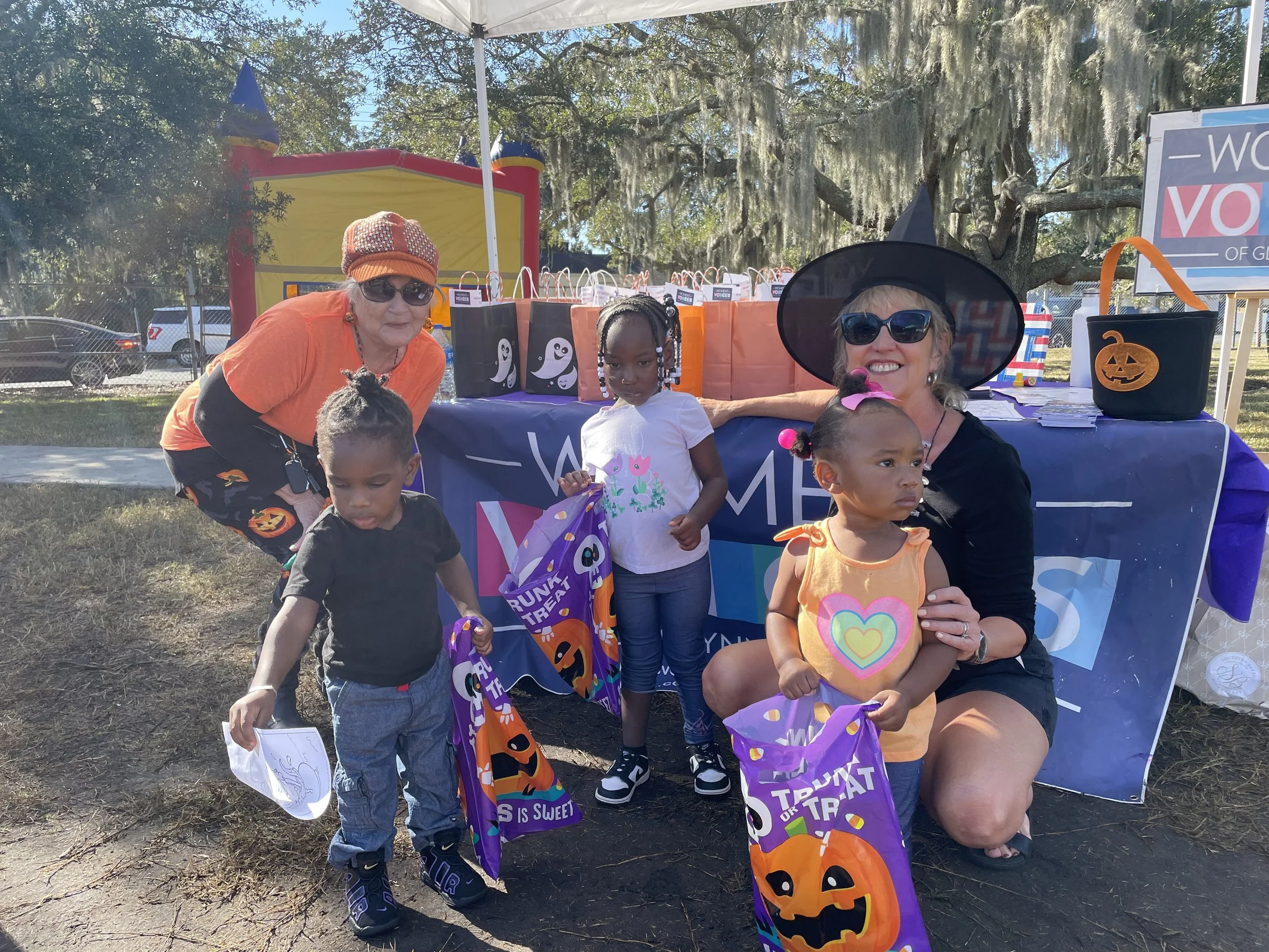 A group of children and two women in Halloween costumes standing outside at a booth with Halloween-themed bags on a table. The women are wearing witch and pumpkin costumes, and the children are holding treat bags.
