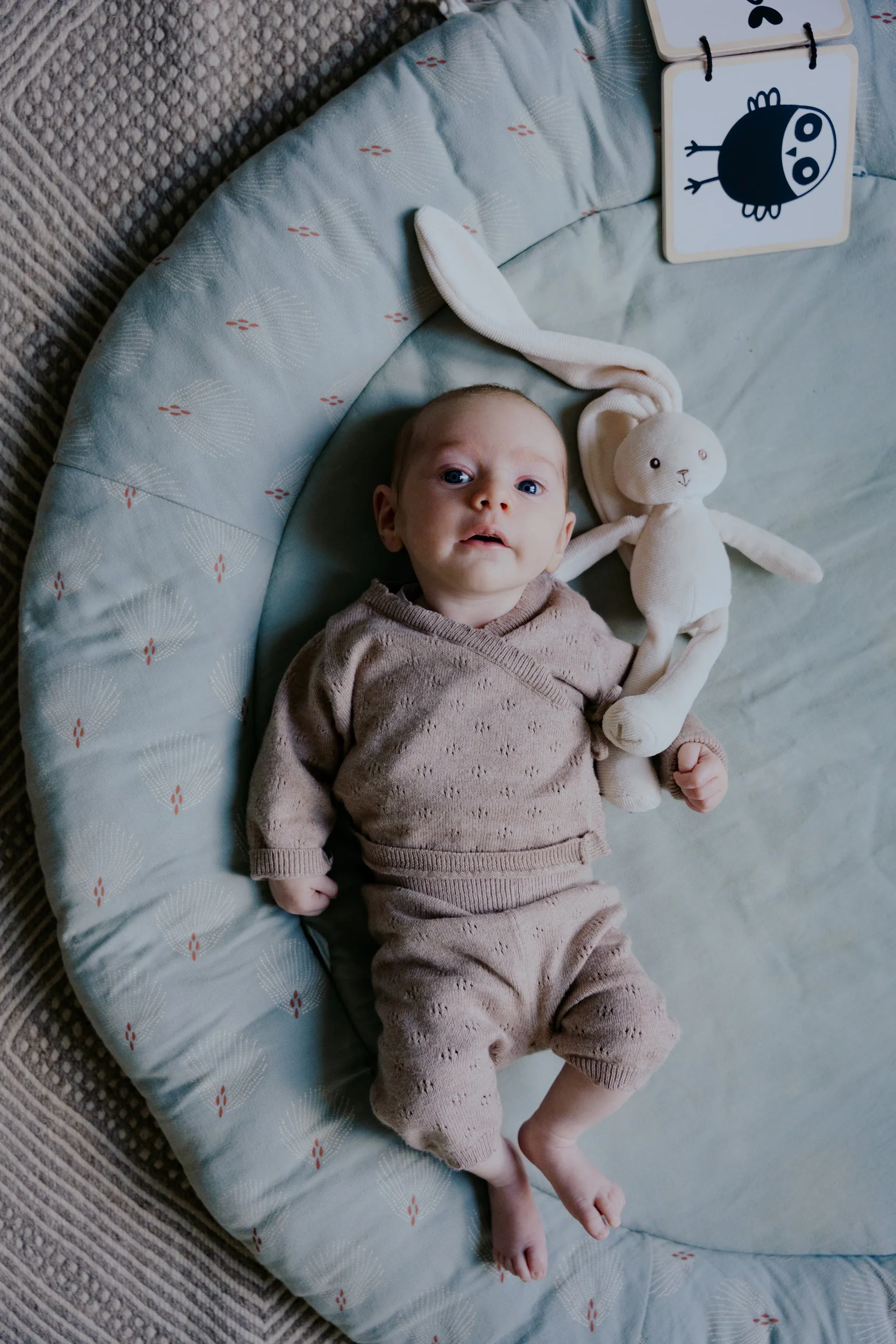 Un bébé allongé dans un lit ou un canapé, portant un body beige. À côté de lui, une peluche en forme de lapin. Le bébé regarde l'appareil photo, avec des draps et un tapis à motifs dans l'arrière-plan.