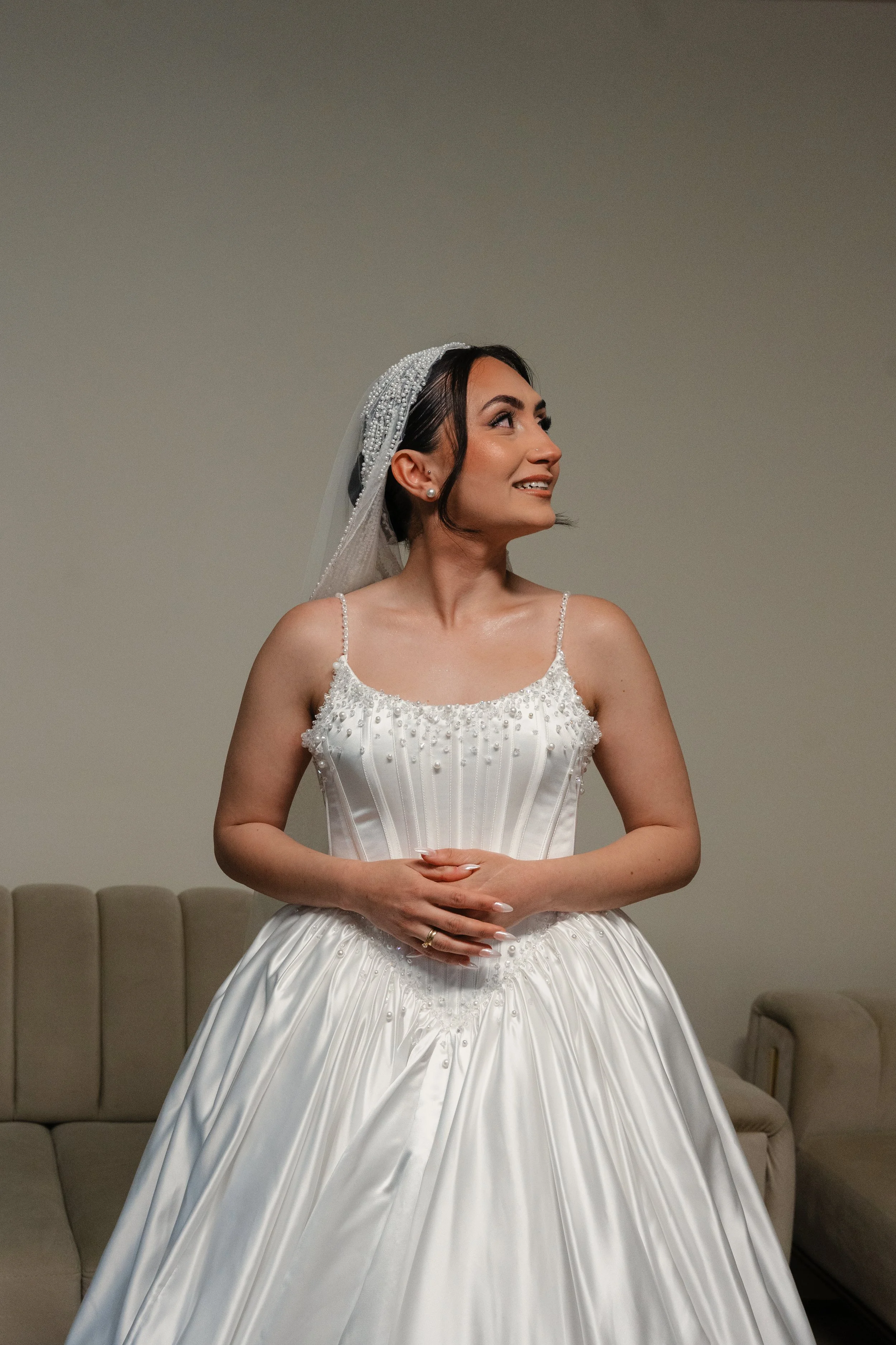 Une femme habillée d'une robe de mariage blanche, avec un voile et des détails perlés, sourit et regarde vers la droite, debout dans une pièce avec des canapés beige en arrière-plan.