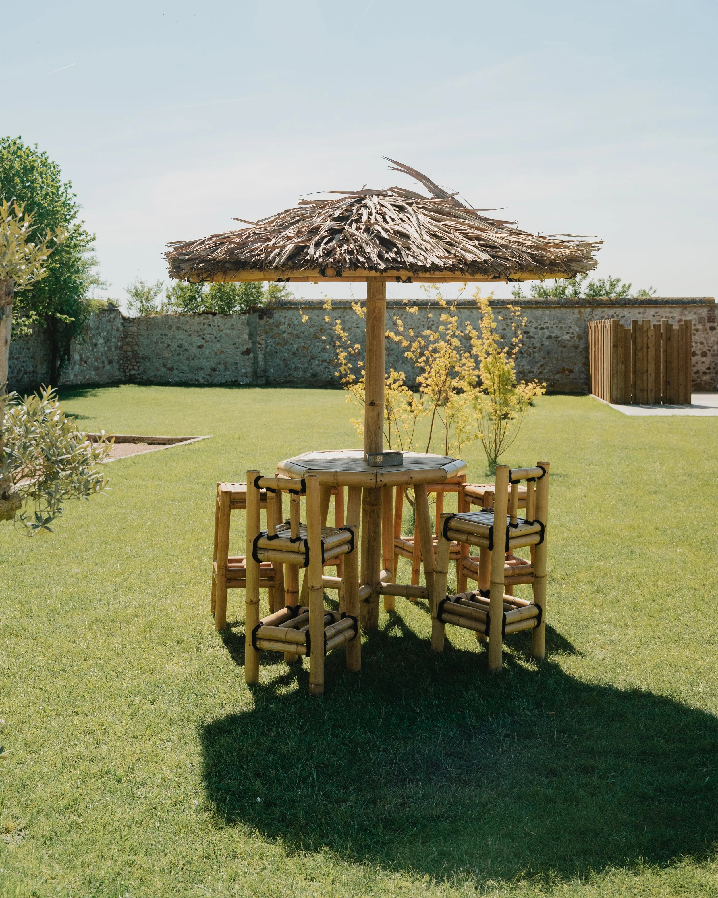 Un parasol en bambou avec un toit en feuilles de palmier, placé sur une pelouse verte, entouré de petits arbustes et d'un mur en pierre en arrière-plan.