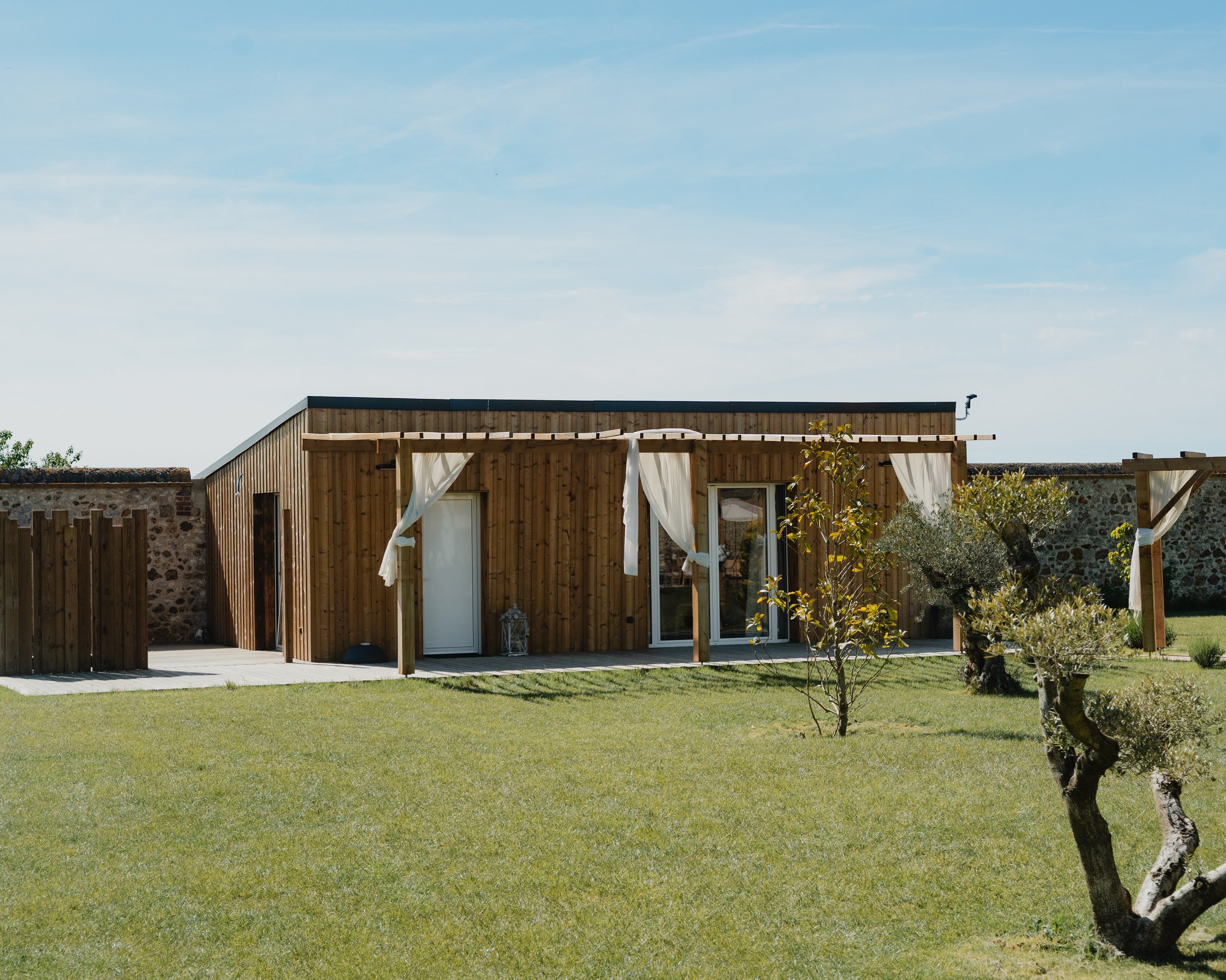 Maison en bois avec terrasse et pergola, jardin avec arbres et pelouse, ciel bleu clair.