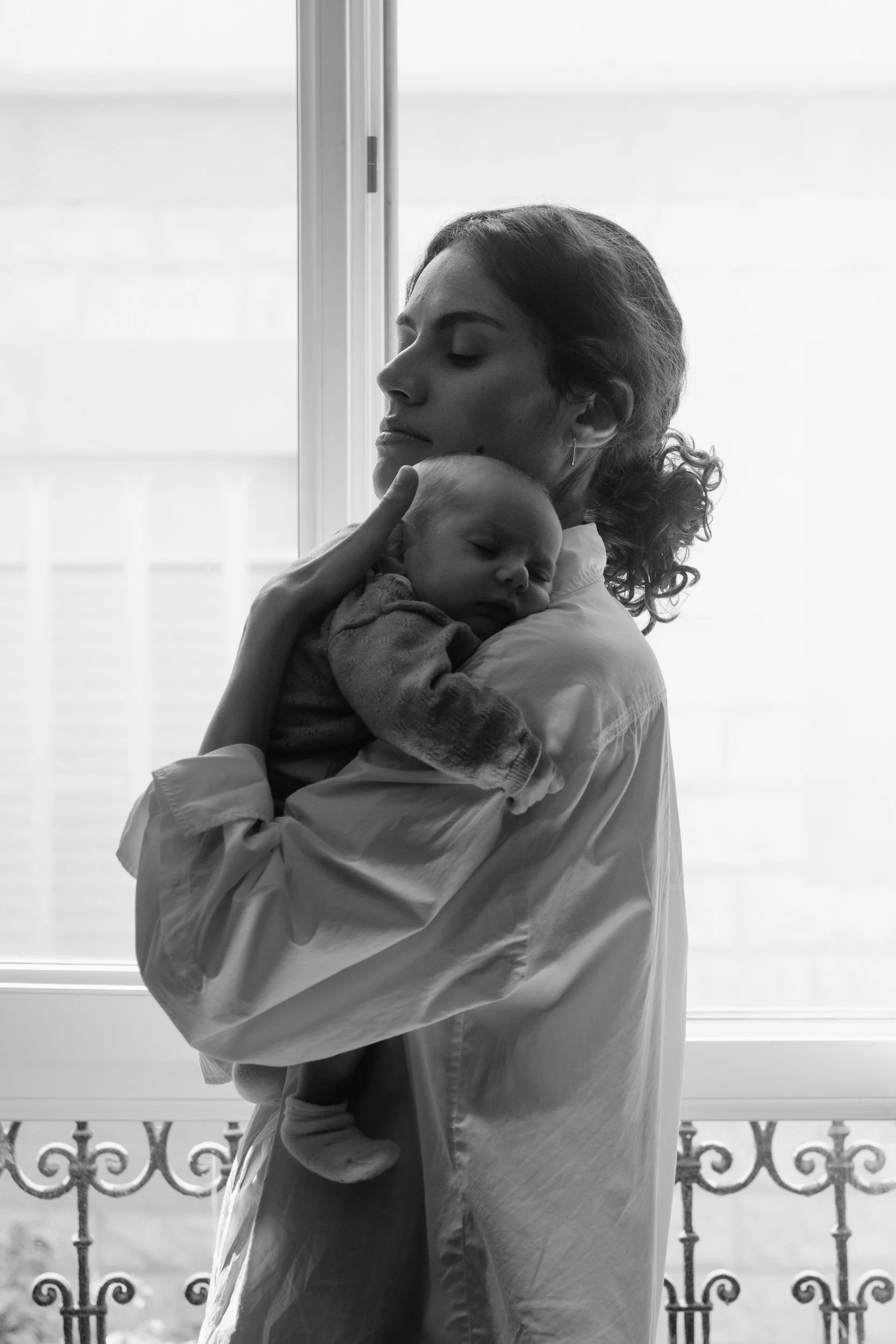 Une femme tient un bébé endormi dans ses bras près d'une fenêtre en noir et blanc.