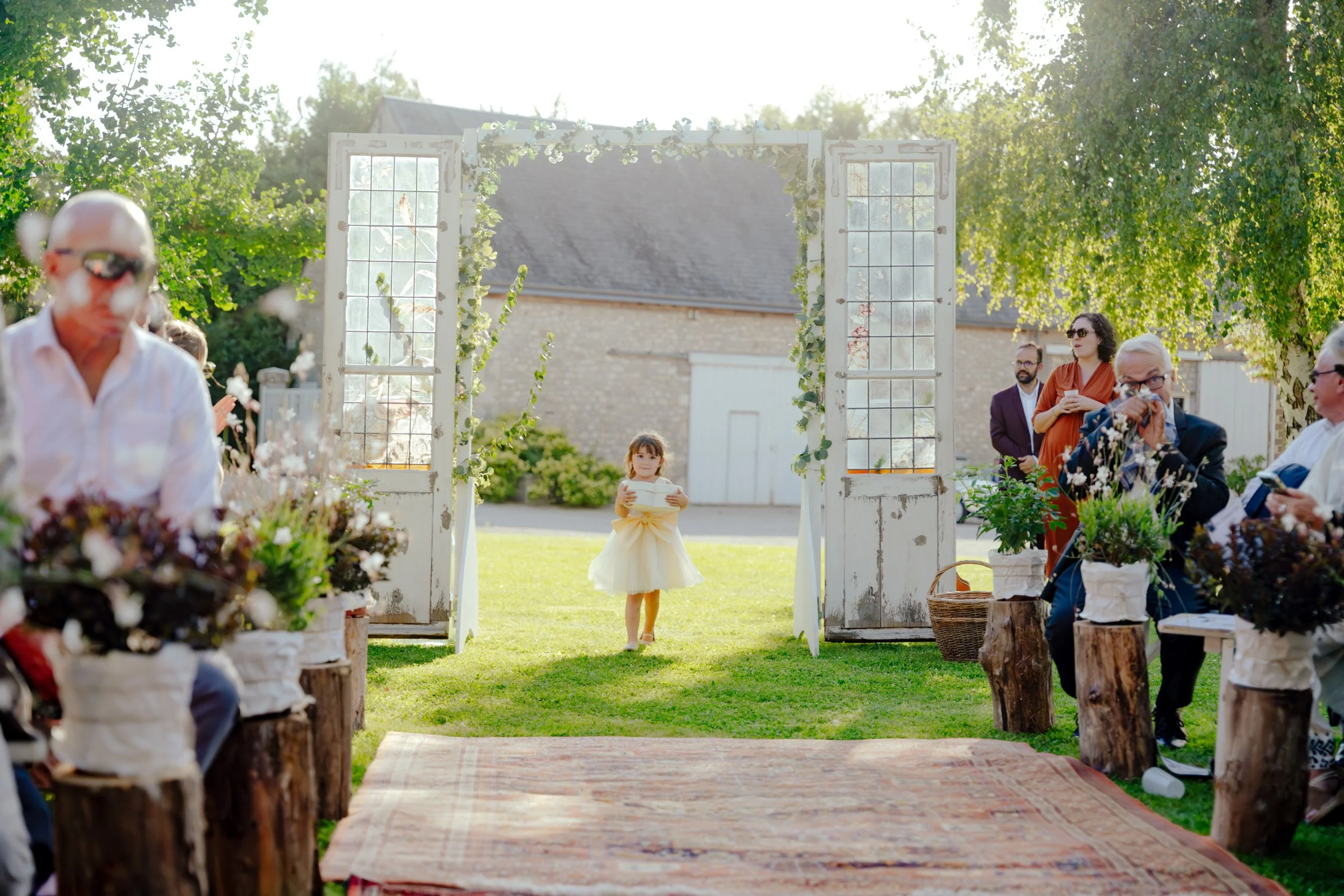 Une jeune fille en robe de fête se tenant dans un cadre décoratif lors d'une cérémonie en plein air. Des invités assistent assis de chaque côté, sous un arbre ombragé, lors d'une journée ensoleillée.