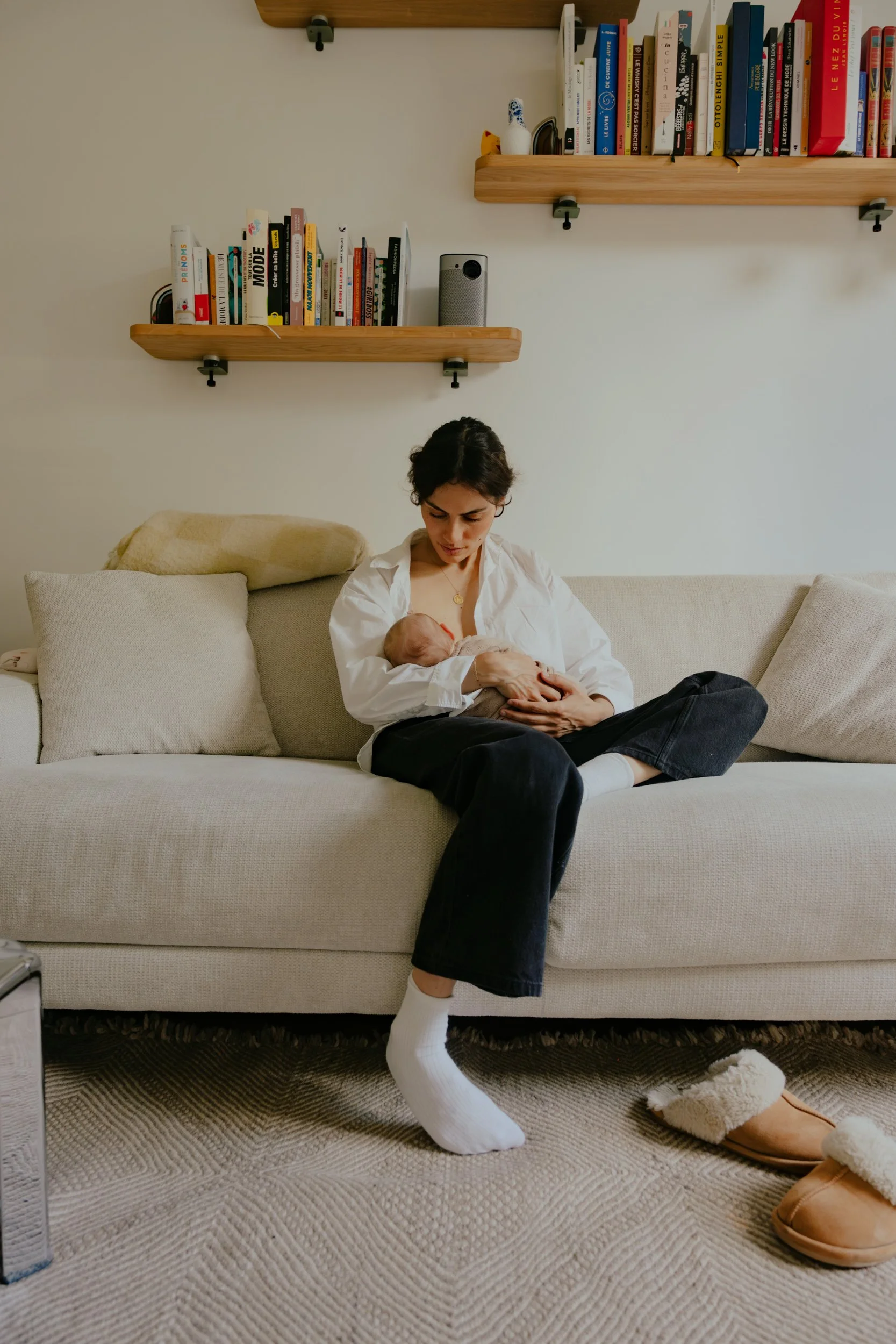 Une femme assise sur un canapé beige, portant un pyjama blanc avec des chaussons douillets et tenant un bébé dans ses bras, dans un salon avec deux étagères en bois remplies de livres derrière elle.